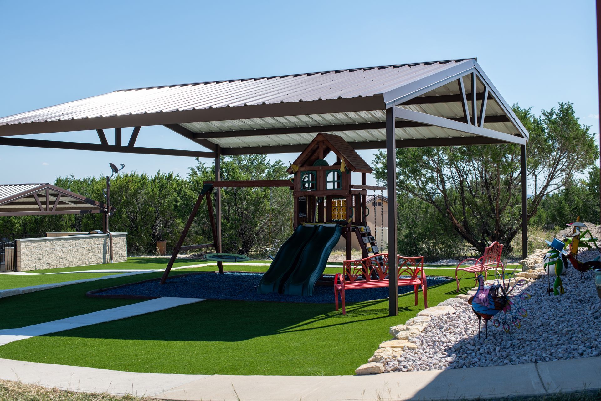 A children's wooden playset with a slide and swings under a metal-roofed pavilion on a green lawn.