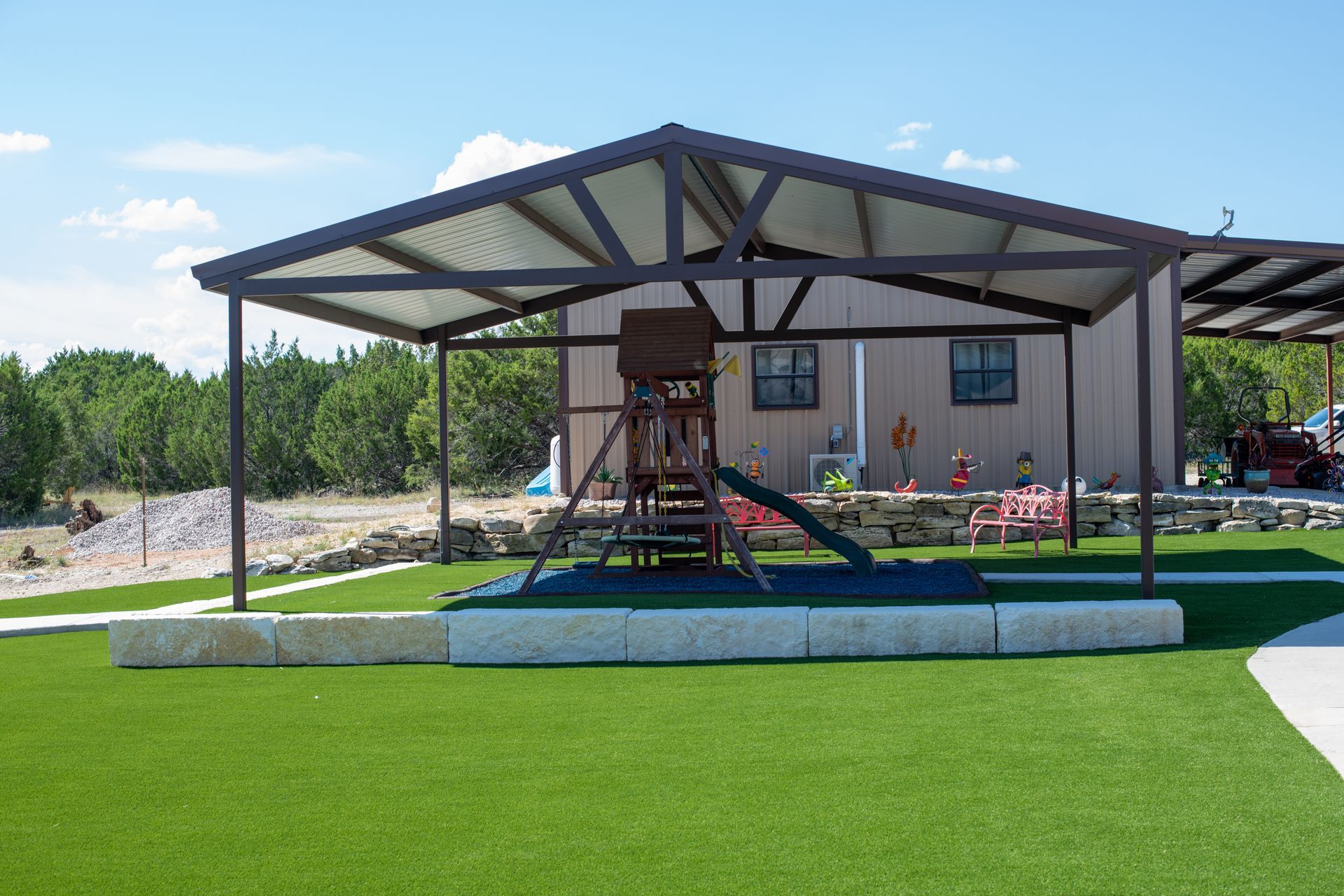 A playground set with a slide sits under a metal canopy structure on a green lawn with a building in the background.