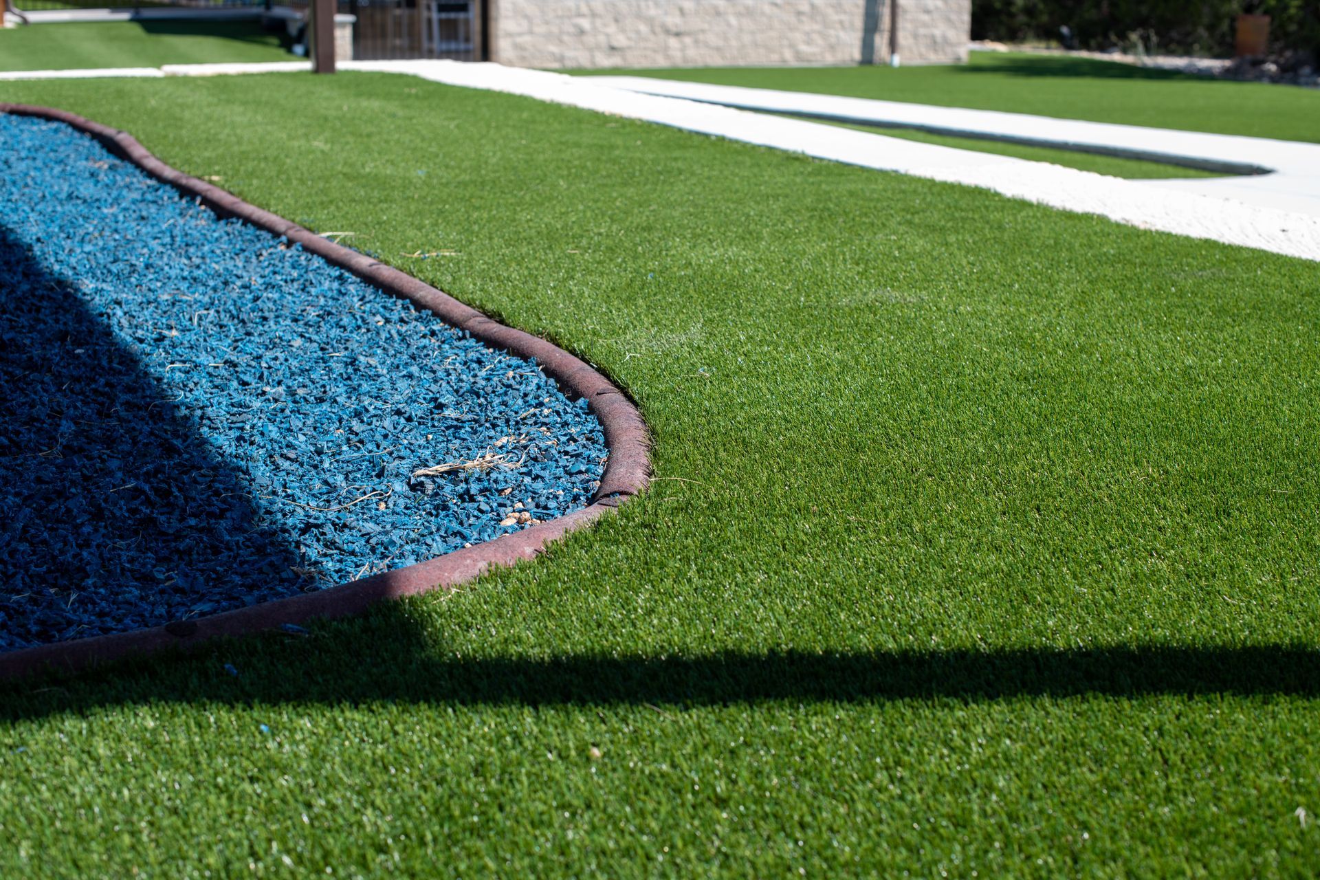 A patch of green artificial turf bordering a curved section of blue landscaping rocks, with a white concrete path nearby.