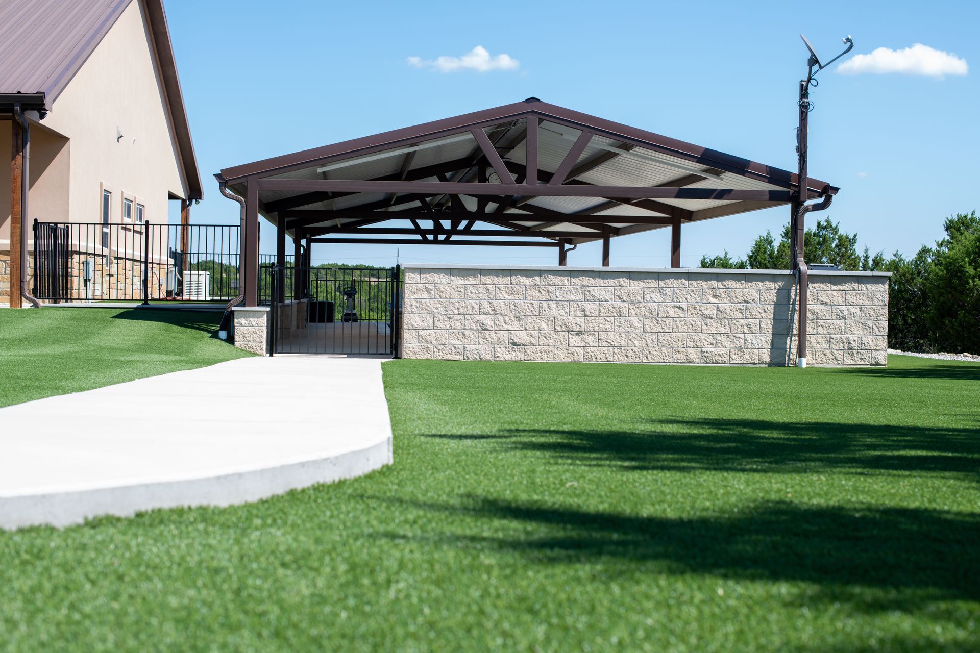 A stone-walled pavilion with a metal roof sits on a green lawn next to a building, connected by a concrete walkway.