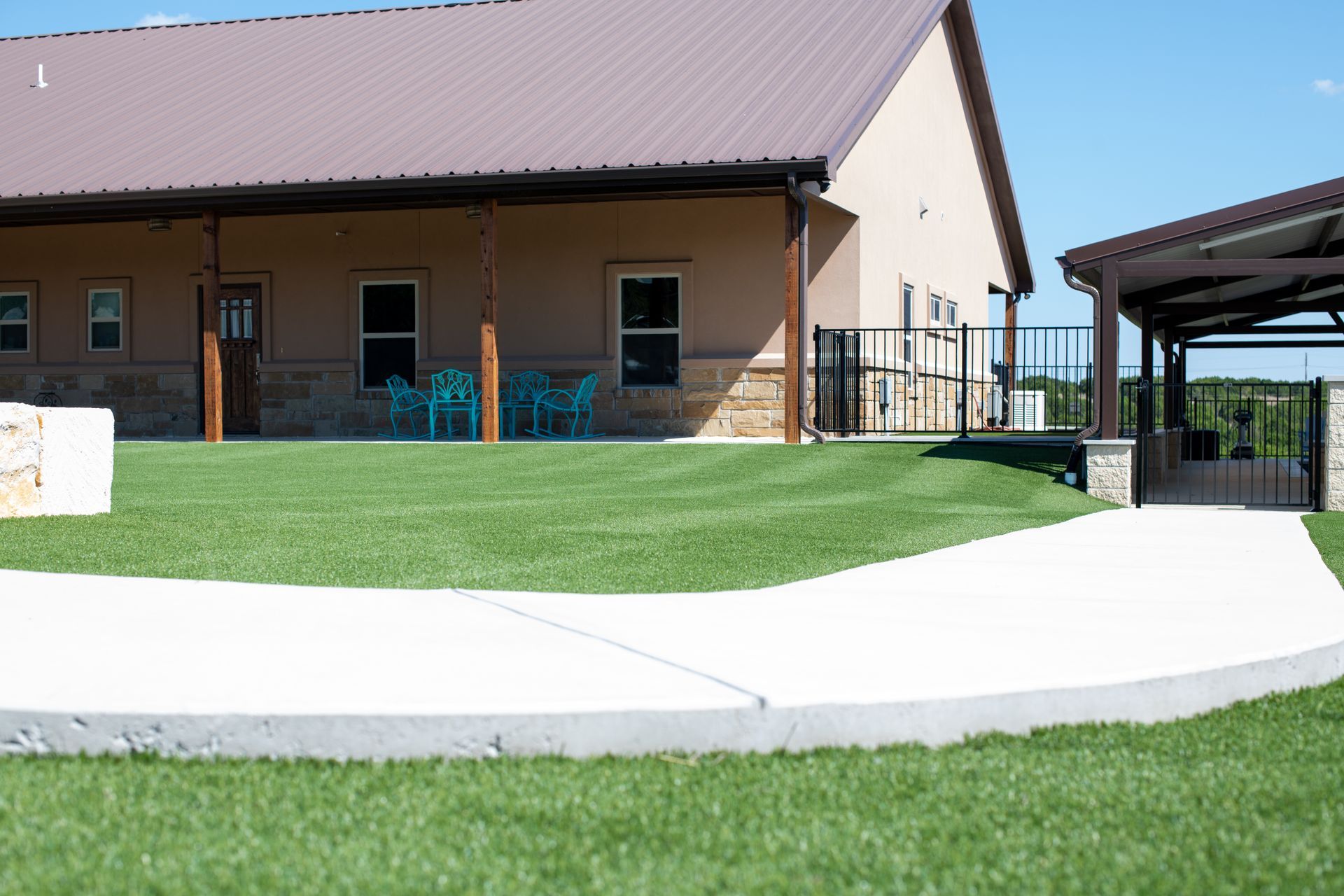 A white concrete walkway leads toward a tan building with a brown metal roof, a stone base, and a small outdoor patio.