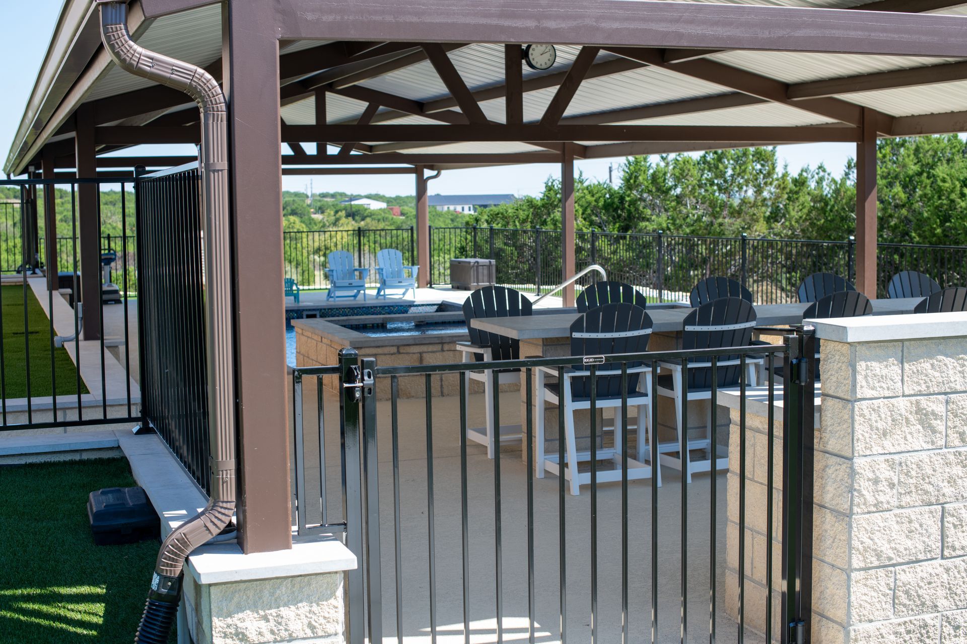 A shaded patio area with outdoor seating, a table, and a metal fence overlooking greenery.