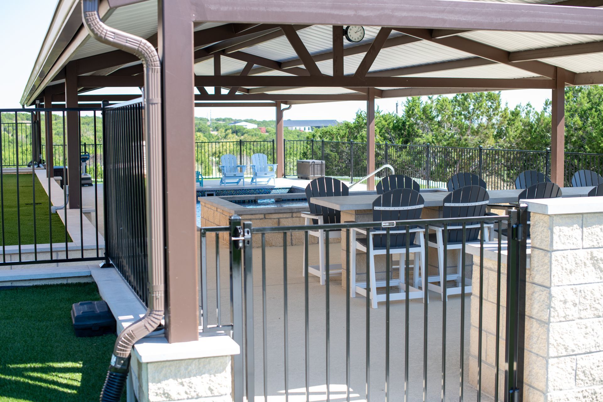 An outdoor patio with a covered seating area, dining table, chairs, and a fence, overlooking a landscaped yard.