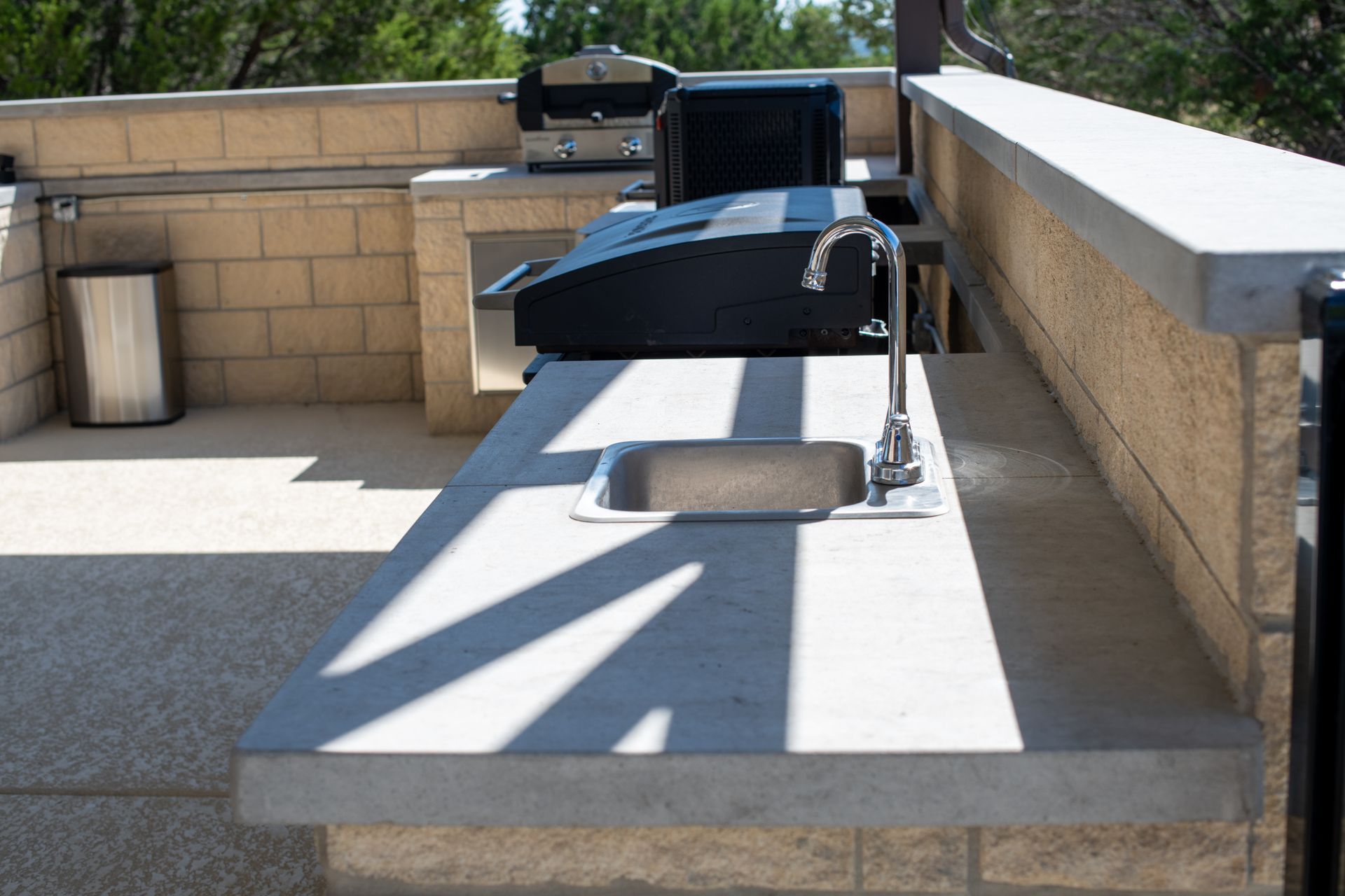 Outdoor kitchen with concrete countertops, a stainless steel sink, and a grill, set against a tan brick wall.