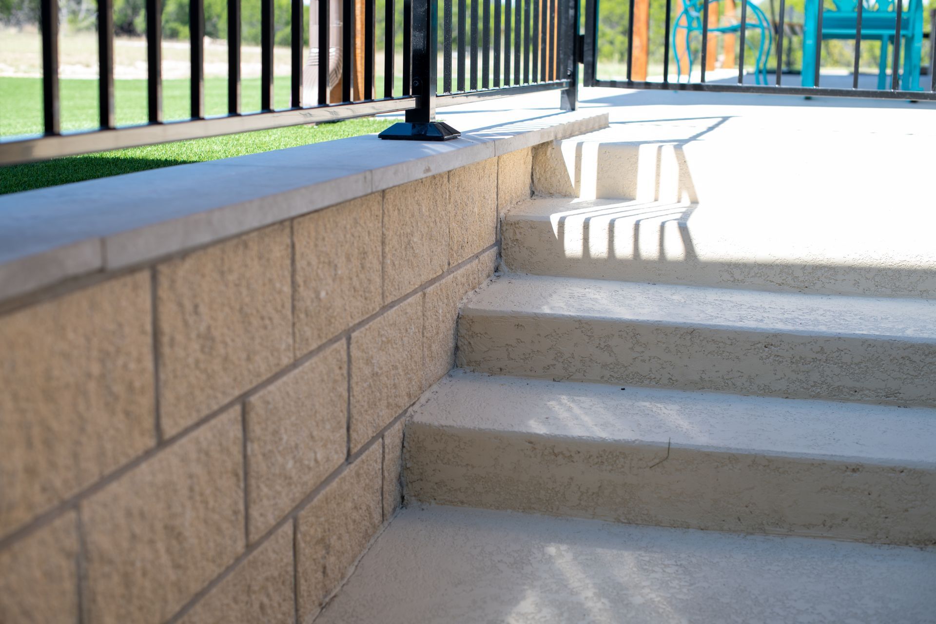Tan stone retaining wall steps leading to a concrete patio with a black metal railing.