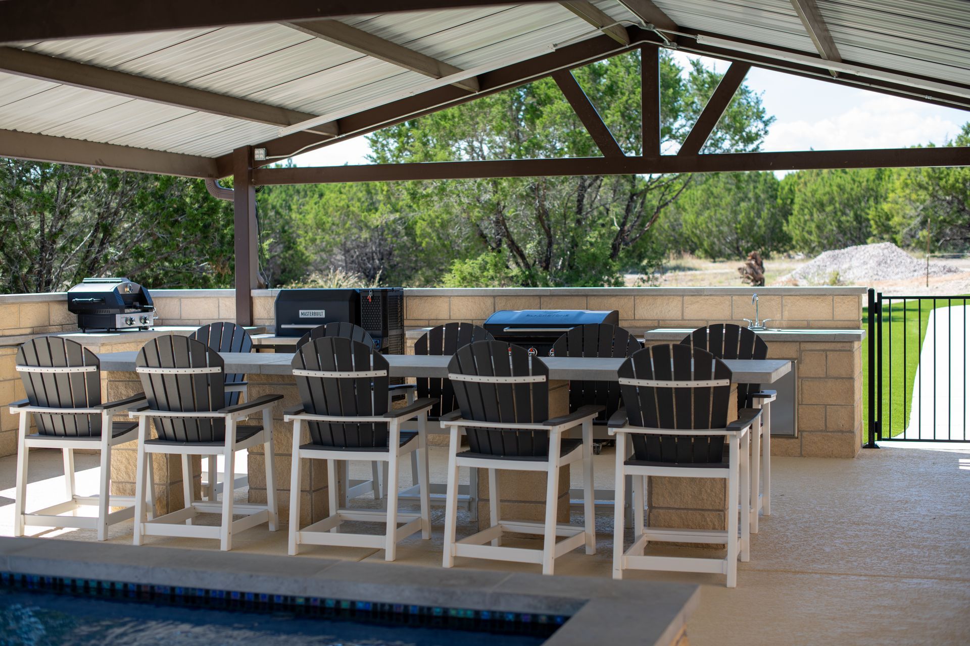 A covered poolside patio with a stone bar, dark chairs, and grills, overlooking trees and a grassy area.