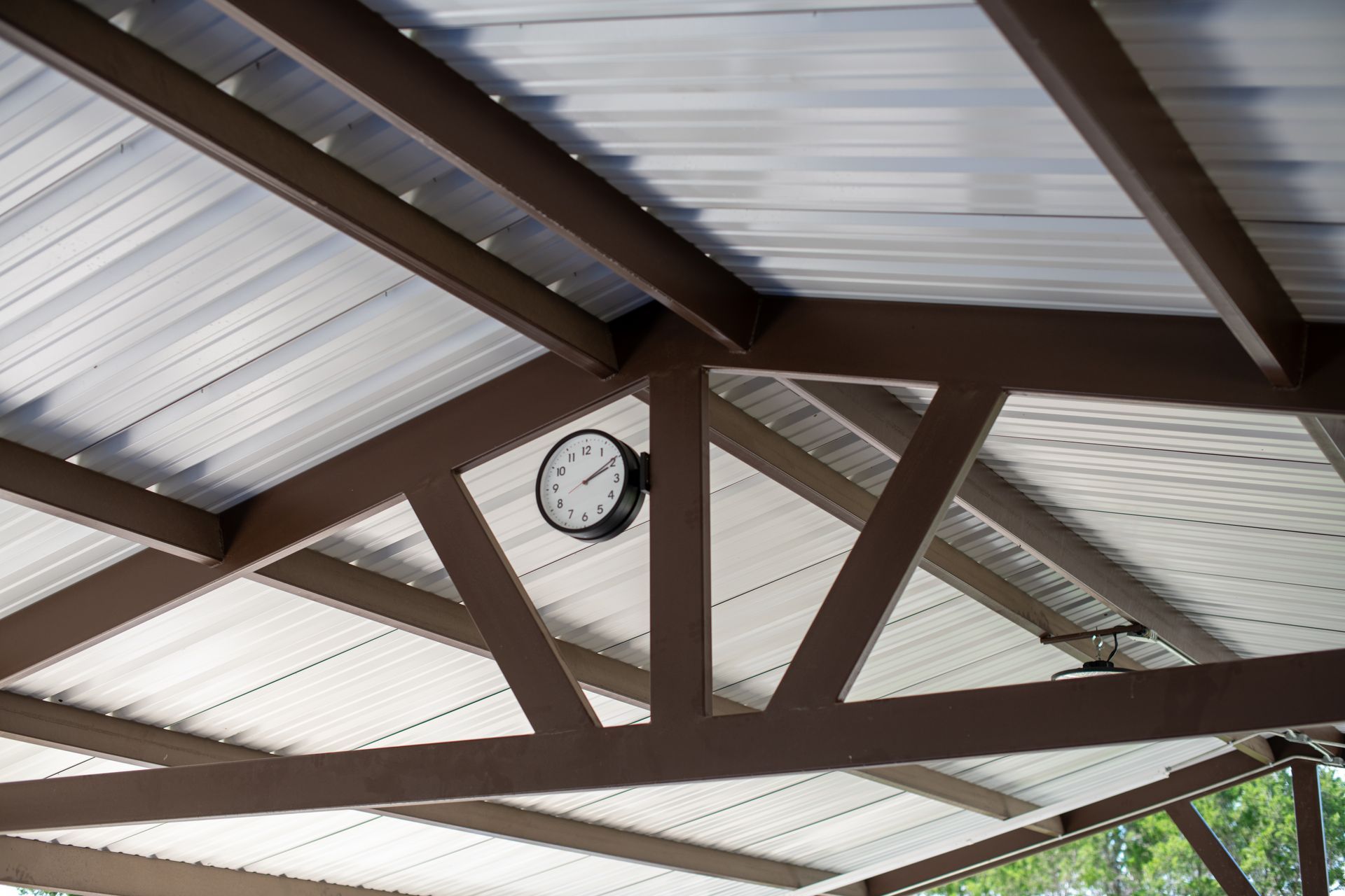 A black wall clock mounted on the brown metal rafters of an outdoor pavilion with a white corrugated roof.