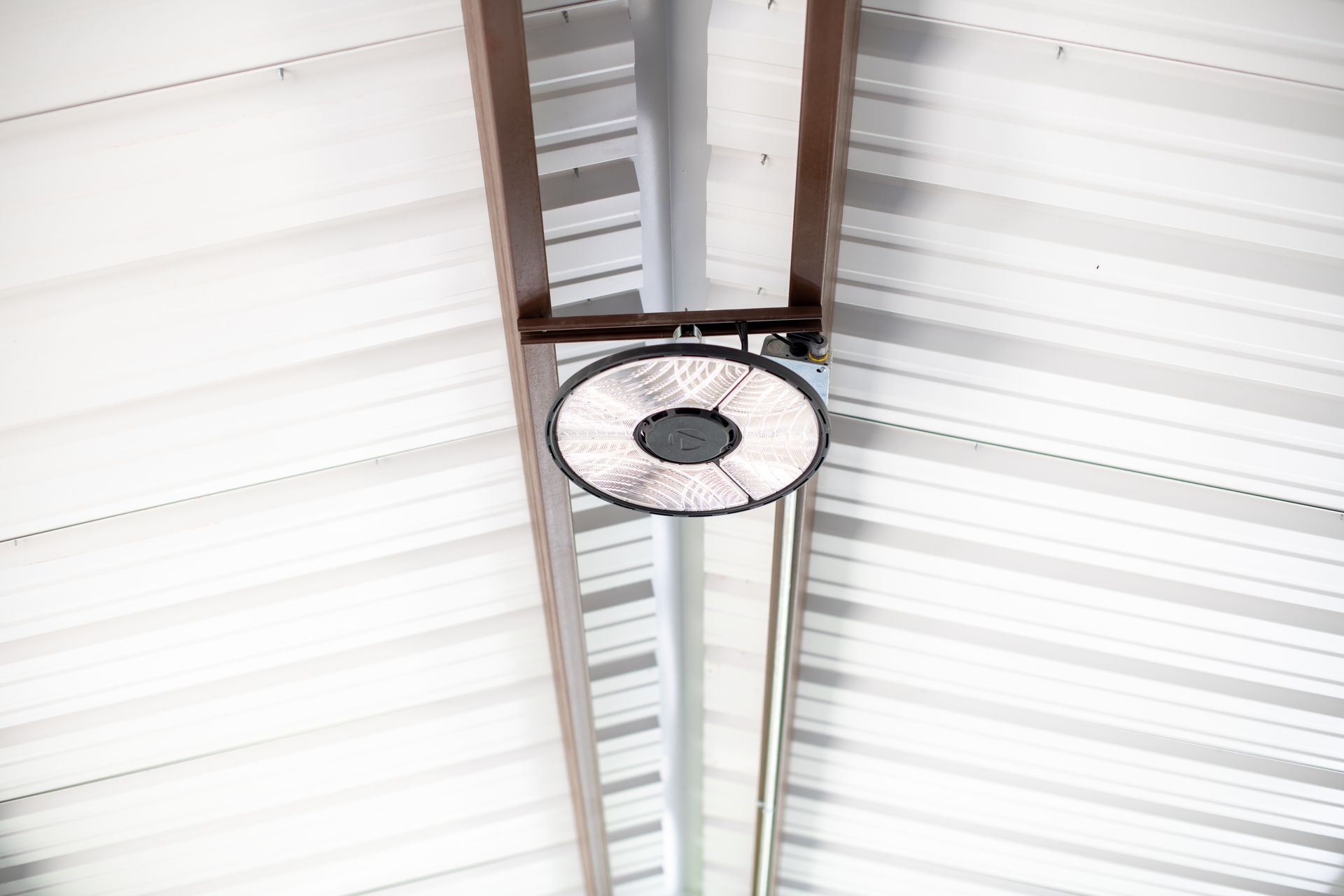 A round LED industrial high-bay light fixture mounted to the metal ceiling beams of a building.