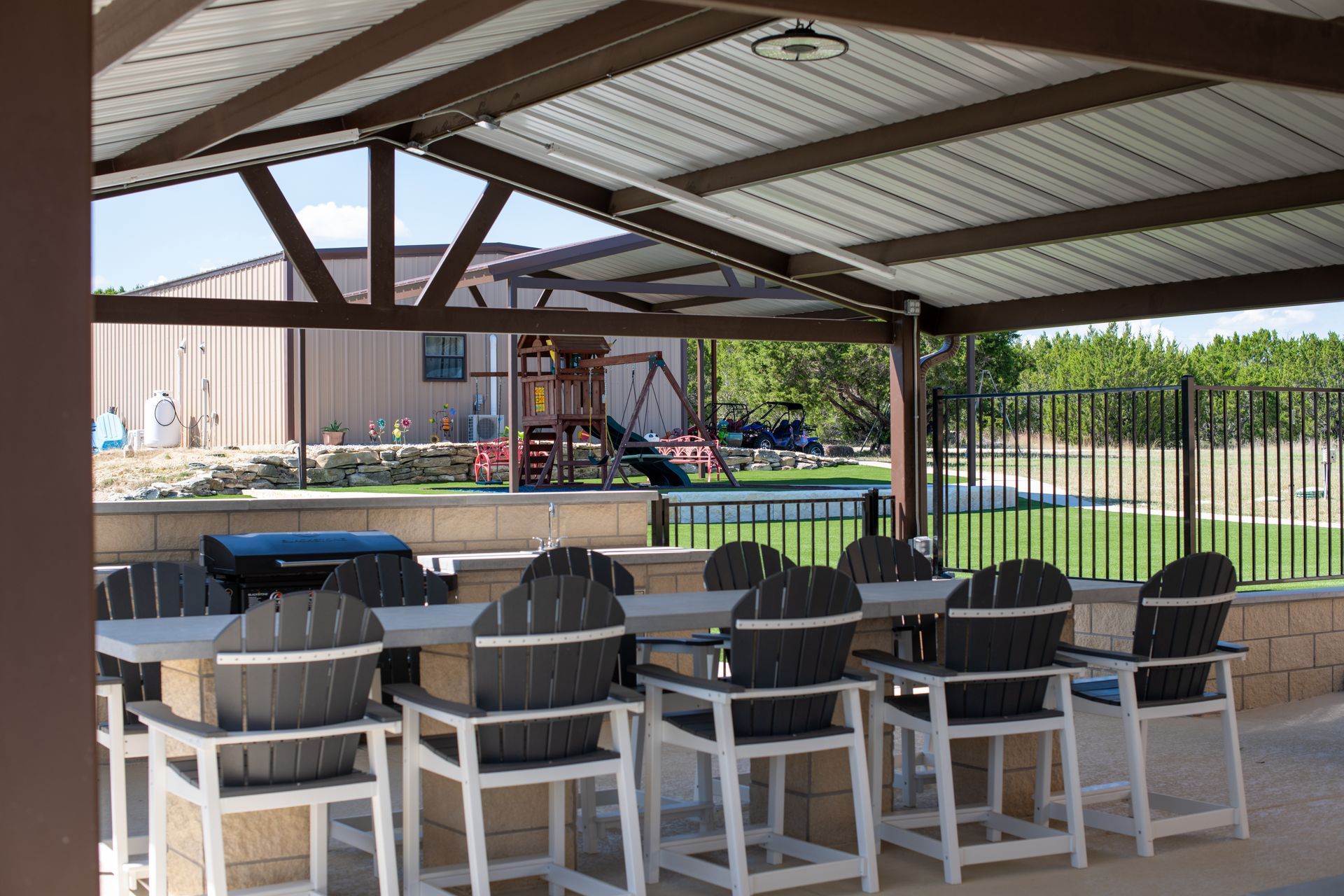A covered outdoor patio with a long concrete bar, several dark-colored chairs, a grill, and a playground in the background.