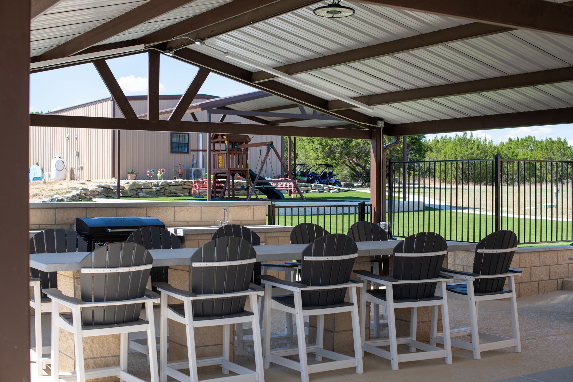 Outdoor kitchen with a bar and chairs under a metal patio roof, overlooking a grassy yard with a playground.