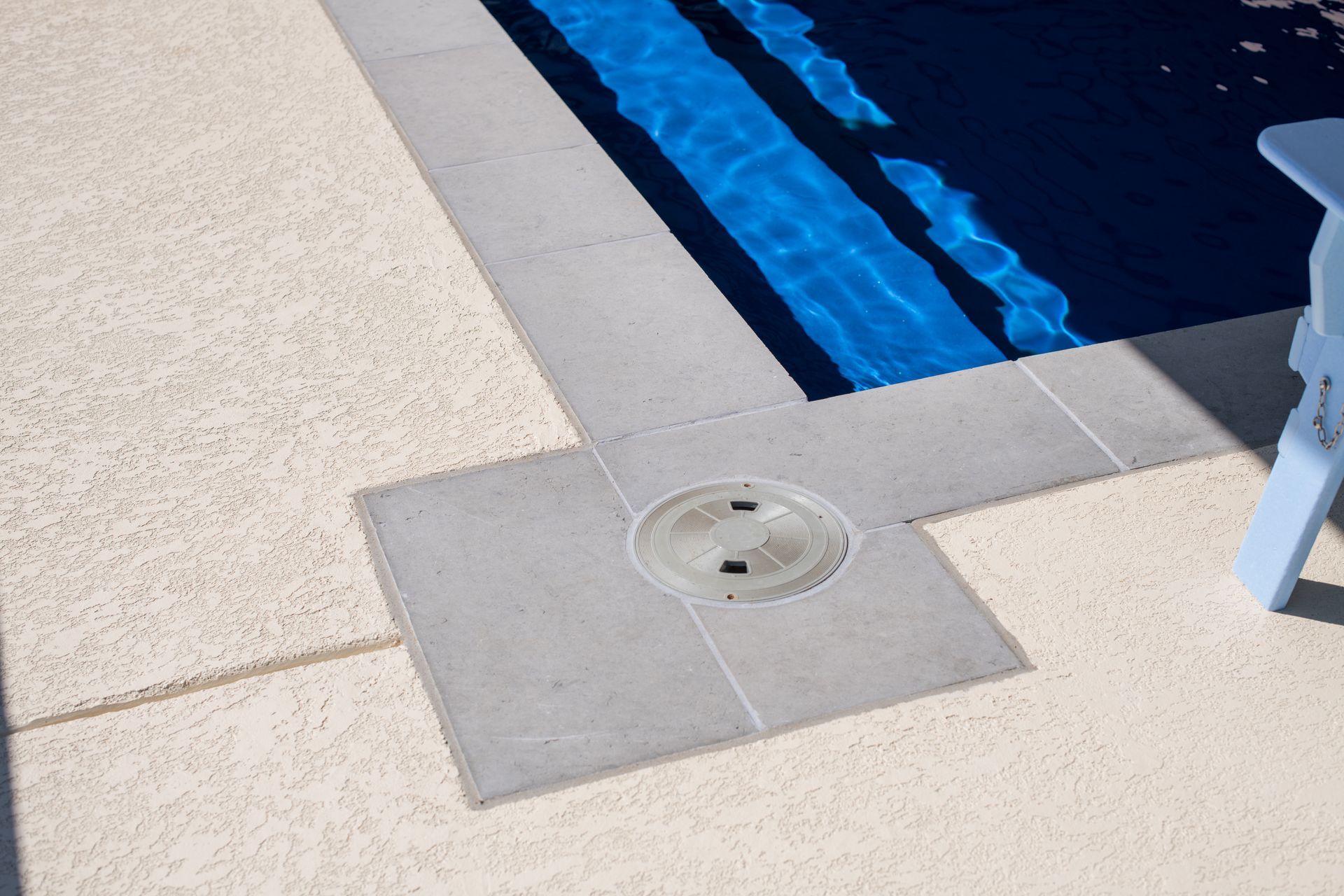 A pool deck featuring a blue-tiled pool edge and a circular drain cover set within gray pavers surrounded by beige concrete.