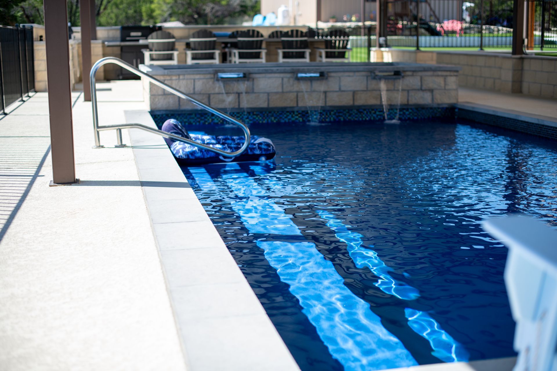 A backyard swimming pool with deep blue water, a stone water feature, a metal handrail, and lounge chairs in the background.