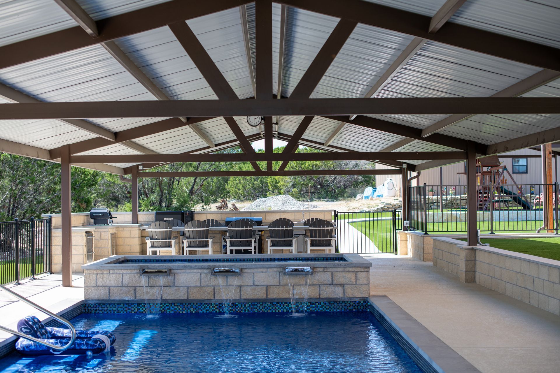 A covered patio with a stone wall, water features, and outdoor seating overlooking a swimming pool and grassy backyard.