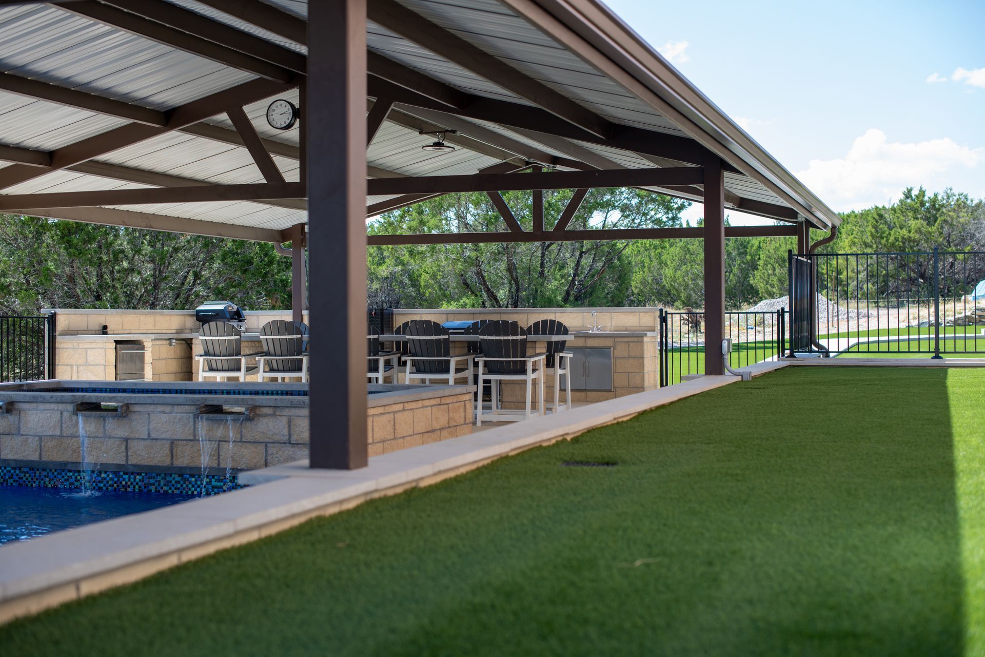 An outdoor patio with a covered kitchen, bar seating, a swimming pool with water features, and a green lawn.