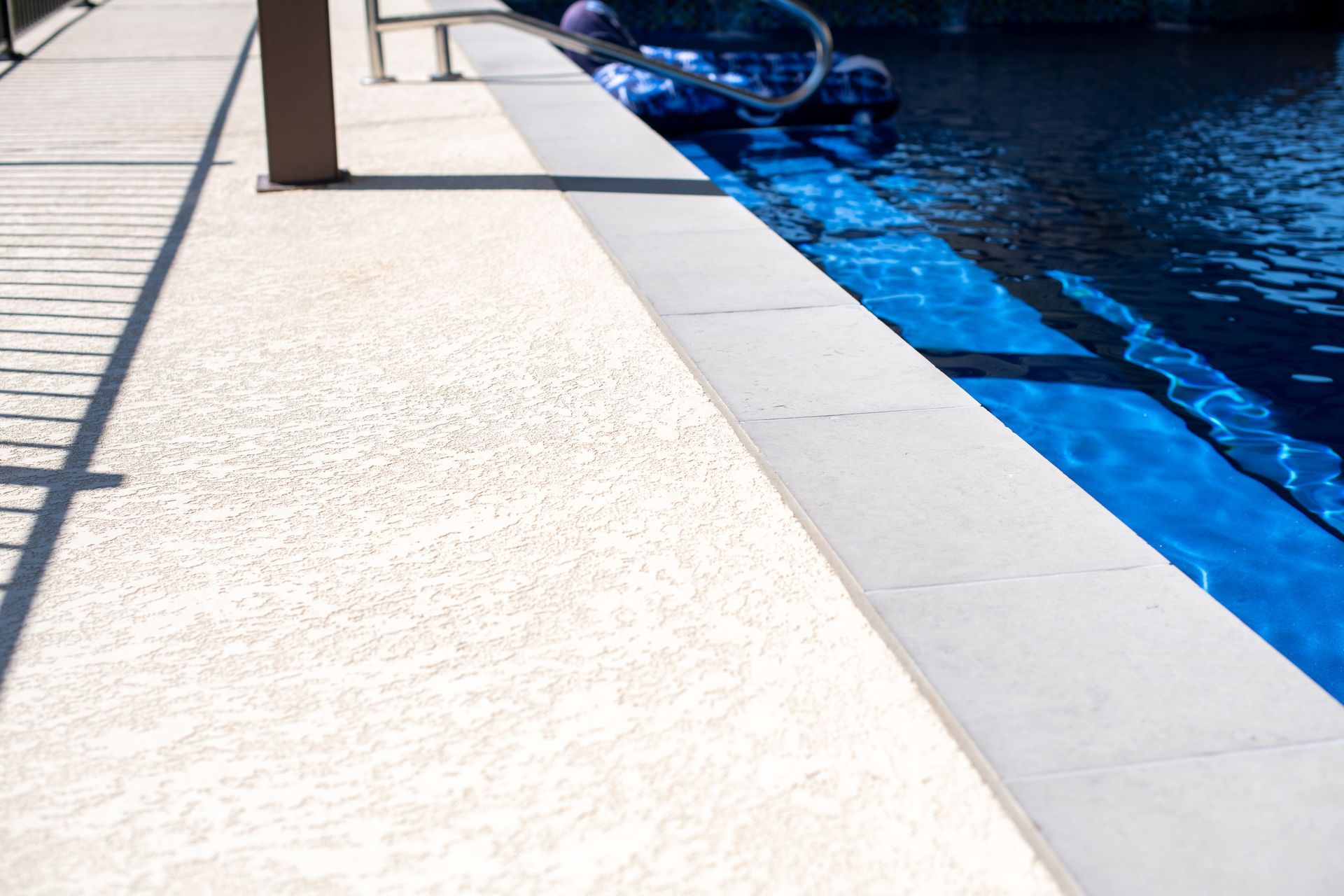 A close-up view of a light-colored, textured pool deck beside a dark blue swimming pool with a metal handrail.