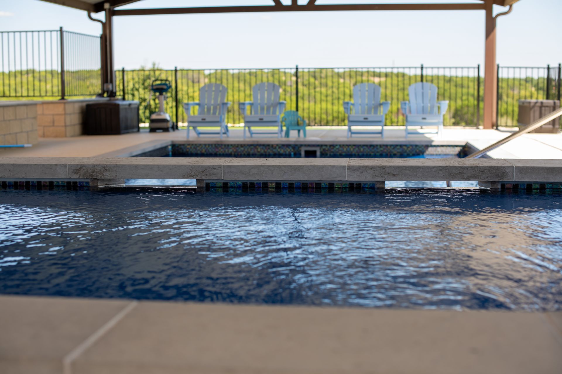 A swimming pool and spa area with four white chairs lined up on a patio under a covered structure with a scenic view.