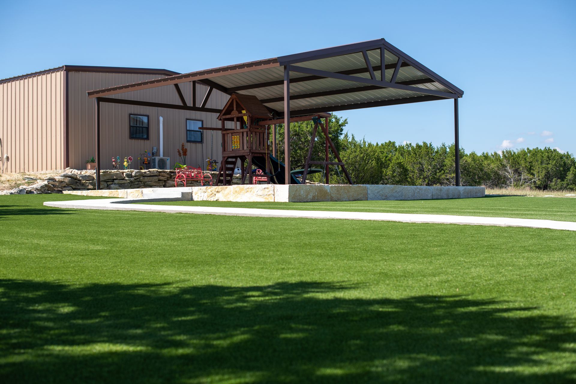 A wooden playground set sits under a metal pavilion on a stone base, next to a beige building and a mowed green lawn.