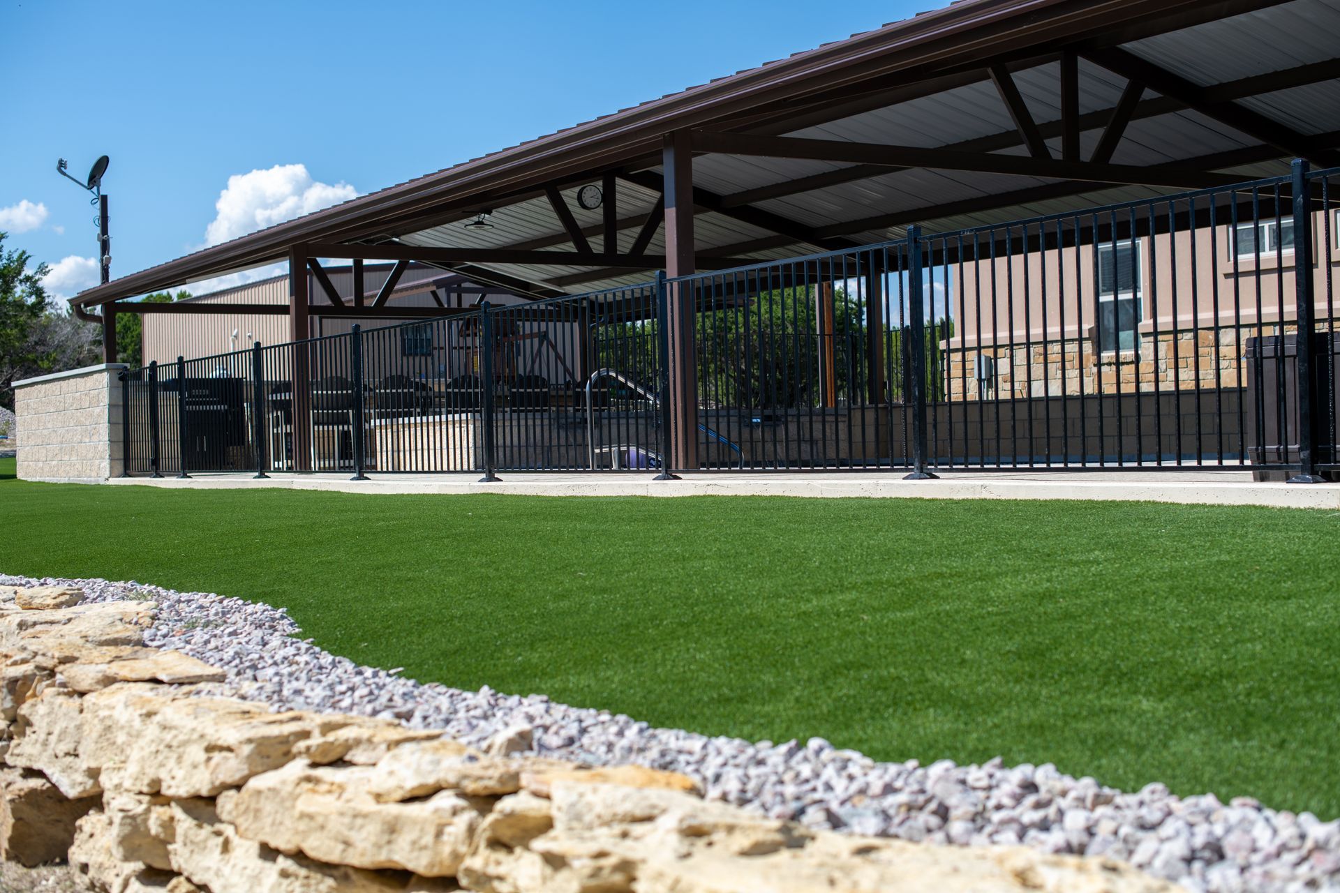 A grassy area next to a stone wall, leading to a covered patio with black metal fencing and a building in the background.