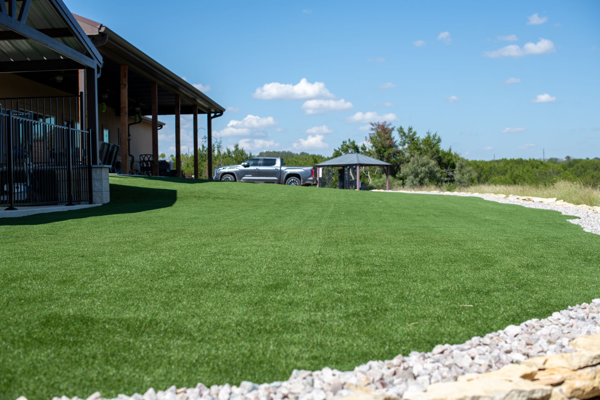 A well-manicured green lawn curves beside a patio building, with a pickup truck and small gazebo visible in the background.