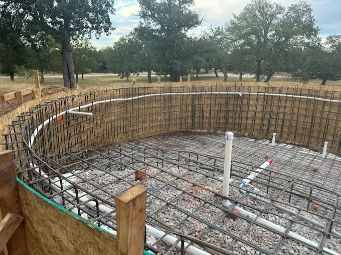 A circular swimming pool structure under construction, featuring a metal rebar cage, plumbing pipes, and wooden forms.