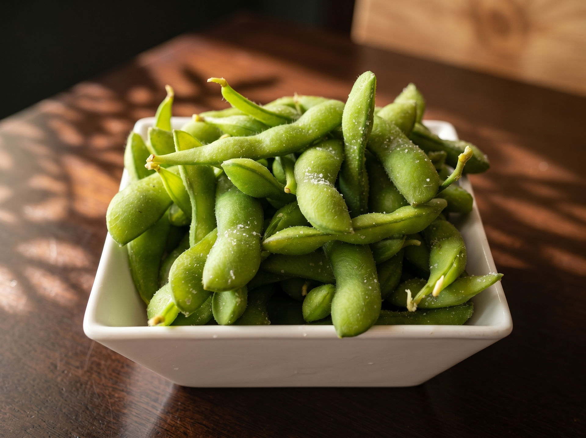 Bowl of green edamame pods on a dark wooden table, lit by warm sunlight