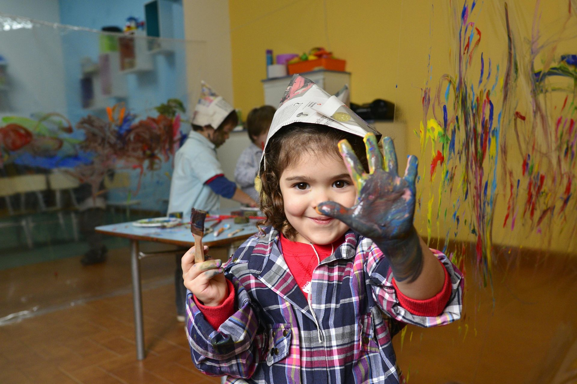 Girl with paint-covered hand smiles, wearing a paper hat, after art class at Kids Creation Stations.