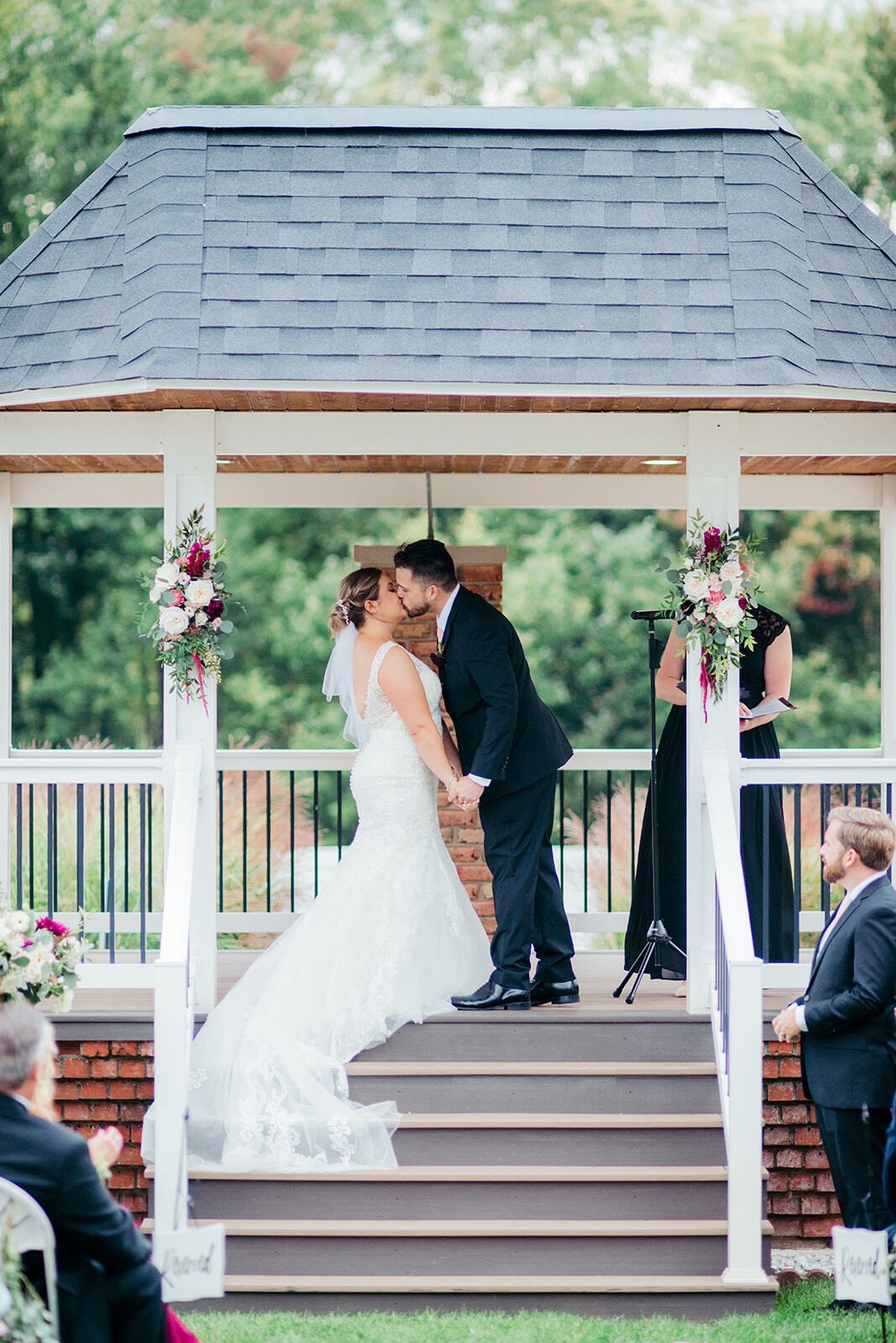Couple kissing at a wedding ceremony under a gazebo with floral decorations.