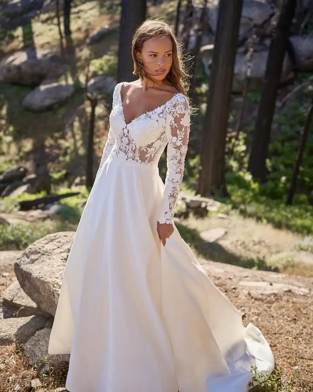 Woman in a white wedding dress with lace sleeves poses outdoors in a sunlit forest.