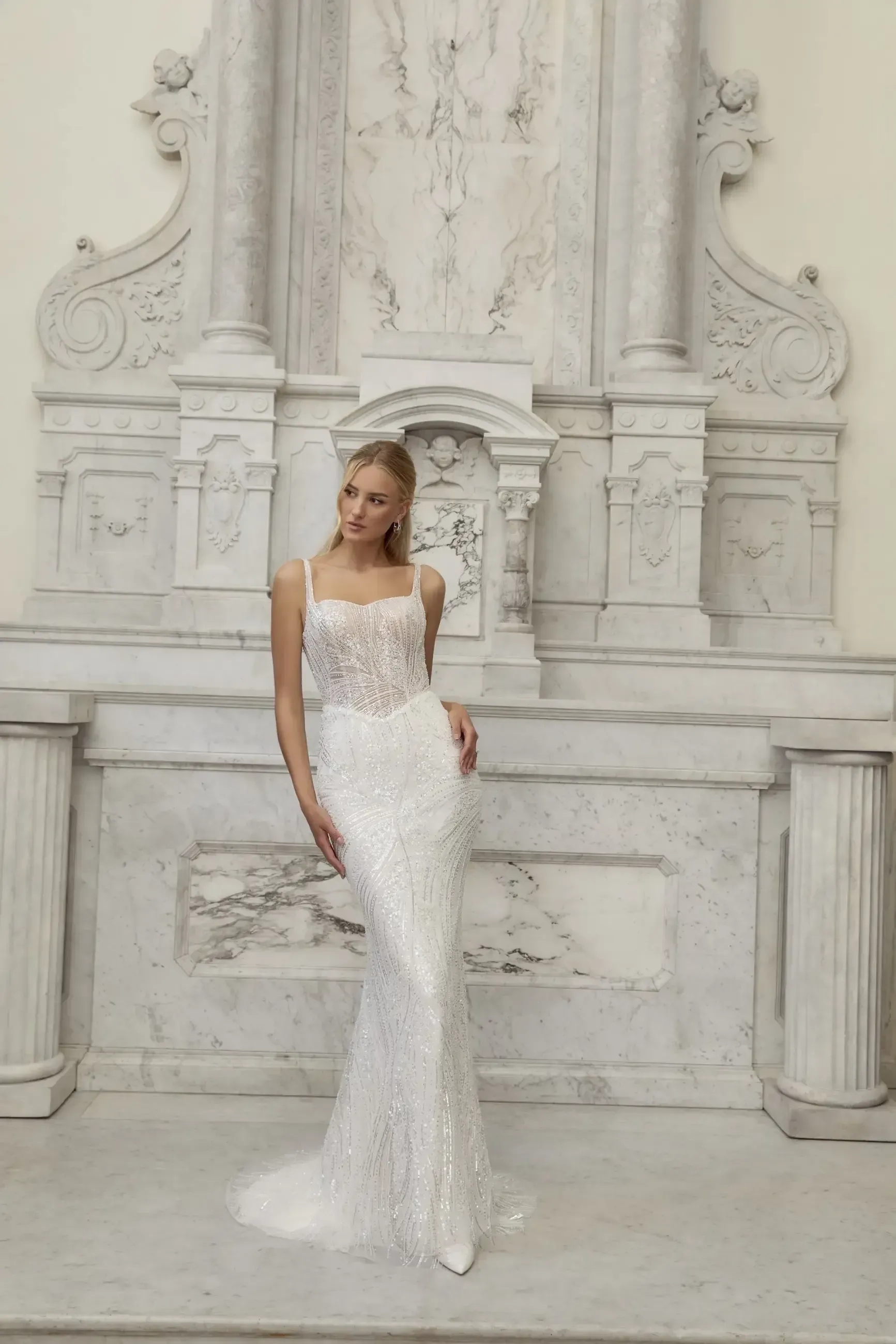 Woman in a sequined wedding dress, posing in front of a decorative, white stone wall.
