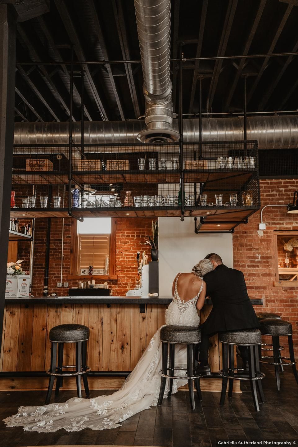 Bride and groom sit at a wooden bar, bride's train flowing. Brick wall and industrial ceiling in the background.