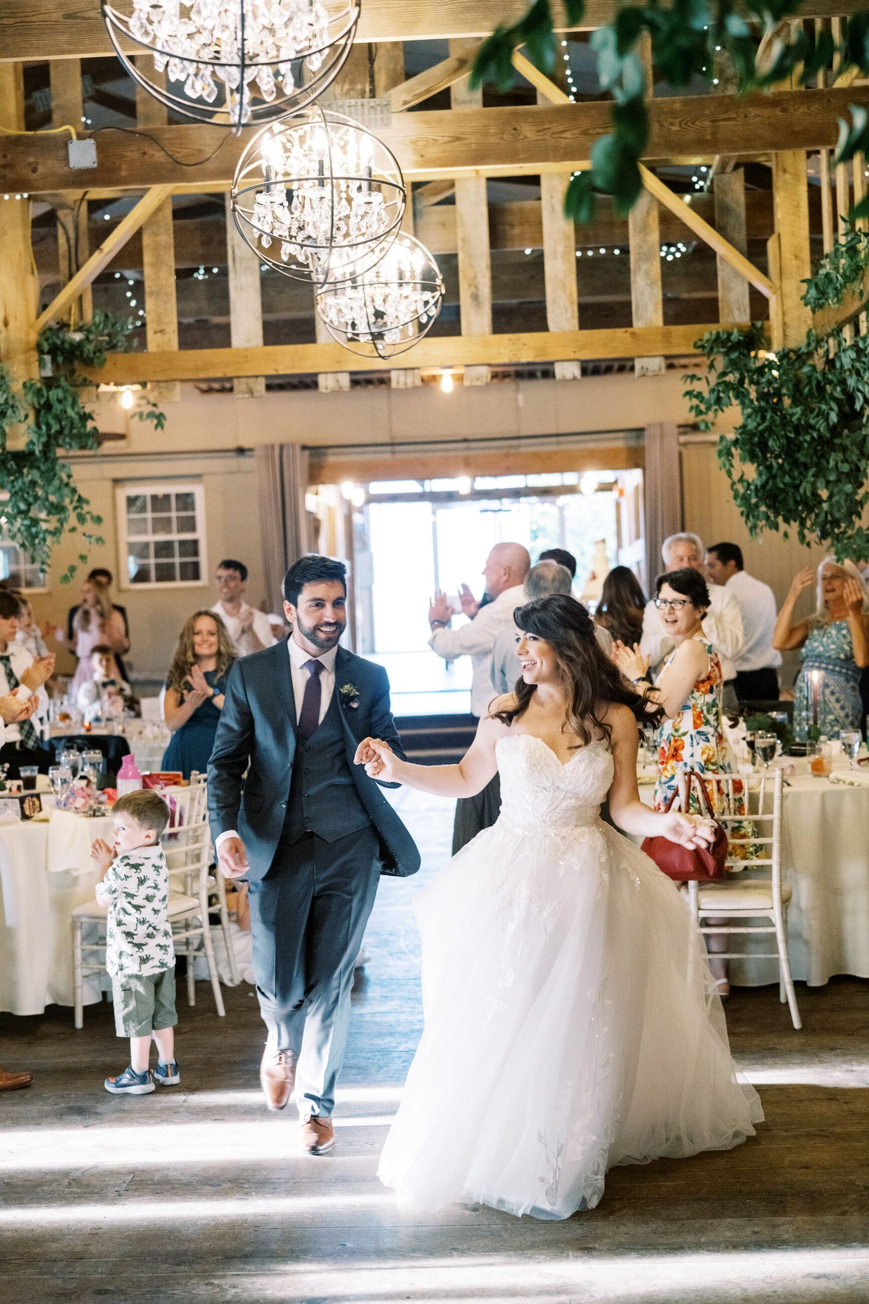 Newlyweds holding hands and entering a reception, guests clapping. Barn setting with chandeliers and tables.