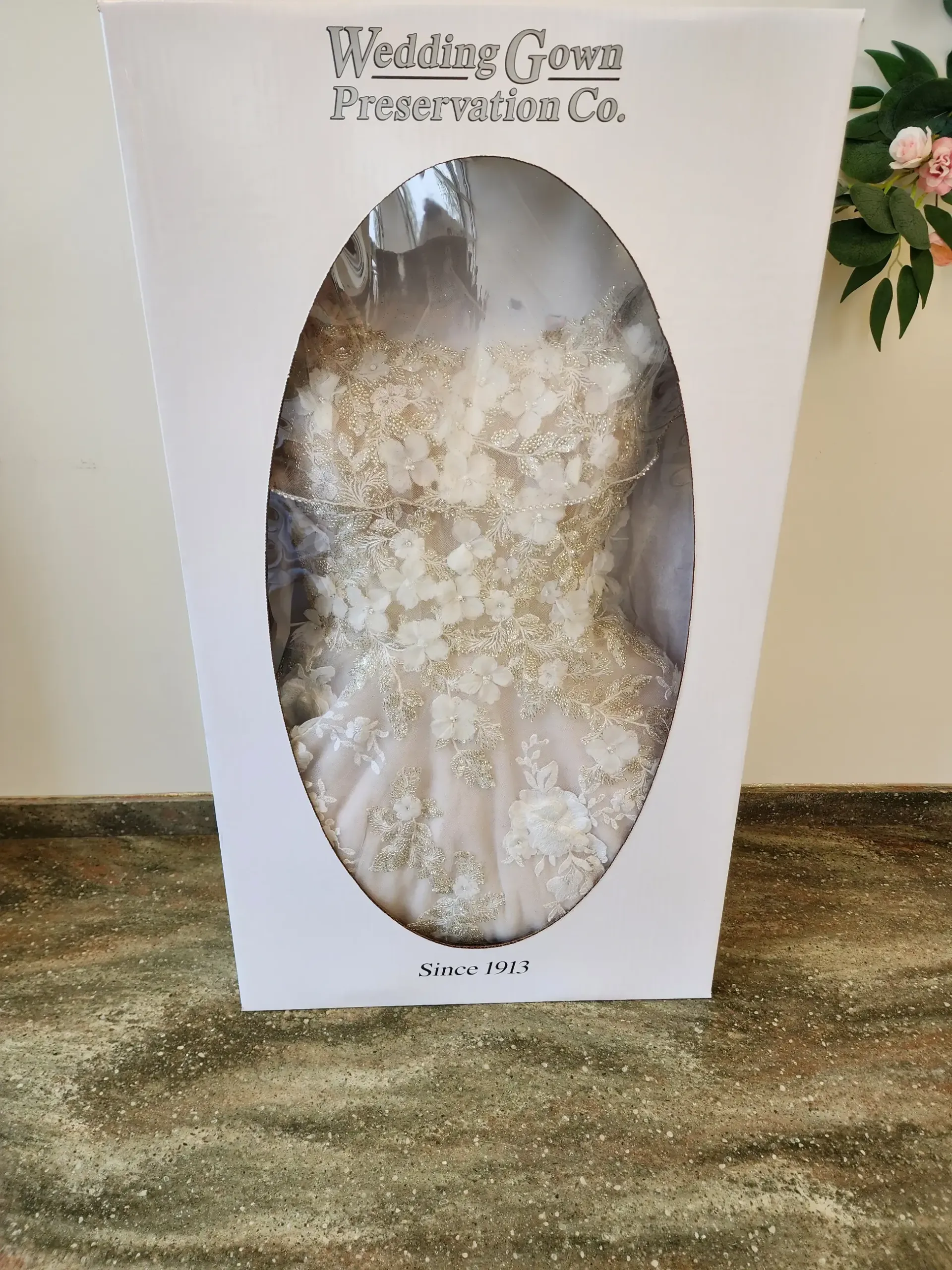 Wedding gown inside a preservation box. Ivory dress with beading visible through the clear oval window.