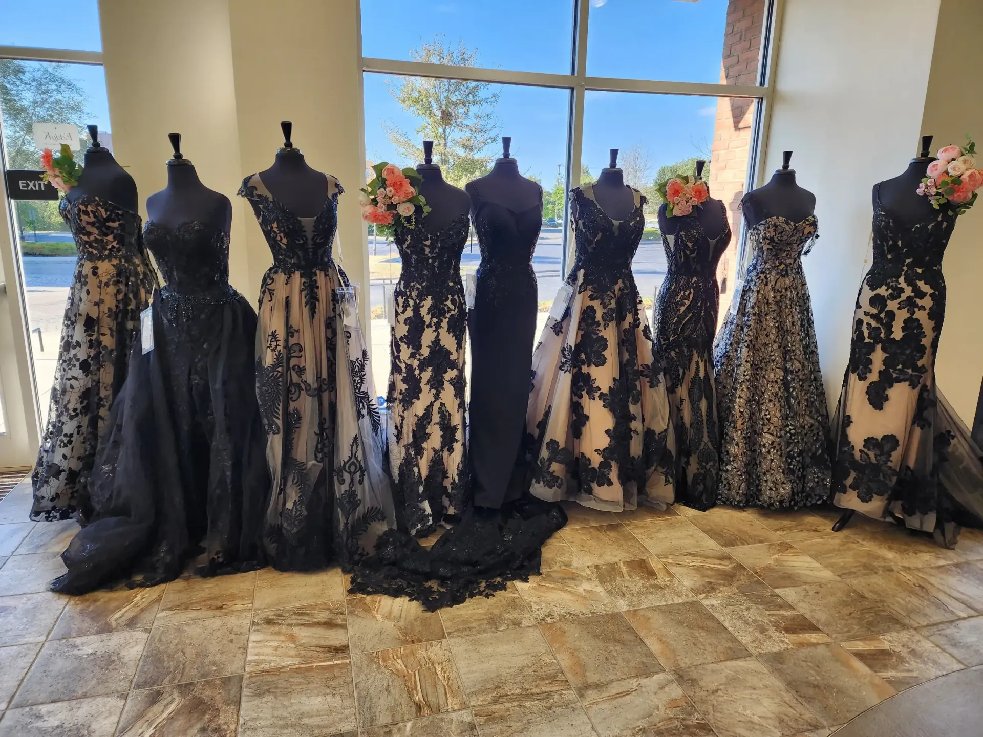 Black gowns with lace detailing displayed on mannequins in a store, near a window.