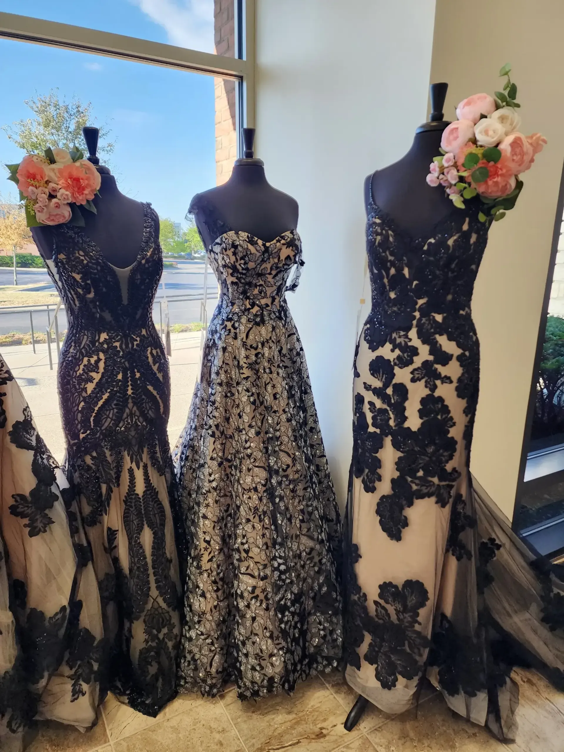 Three formal gowns on mannequins in a window display, featuring black lace detailing over light-colored fabric.