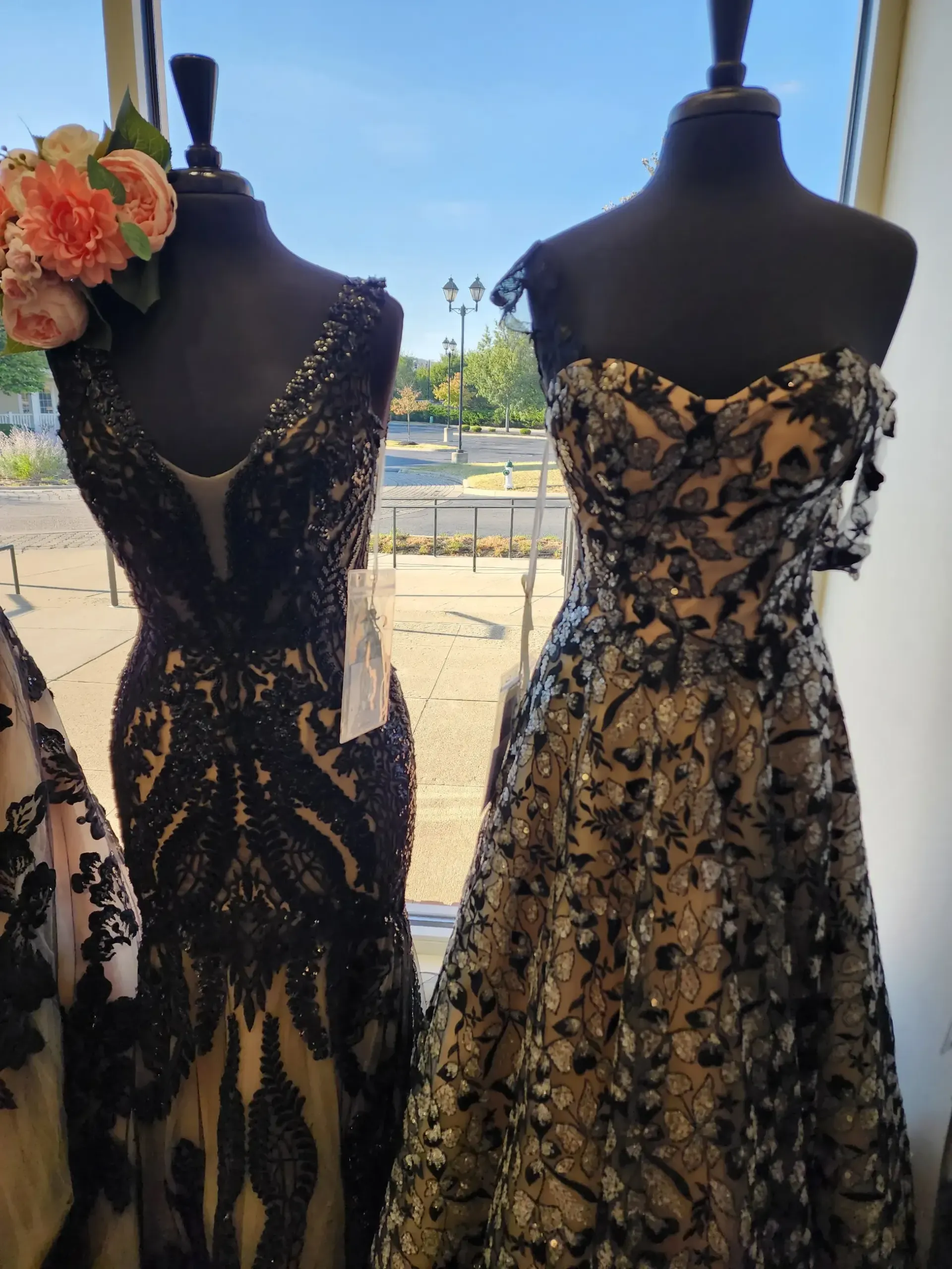 Two formal gowns on mannequins in a window display. One black beaded, one floral design, tan base.