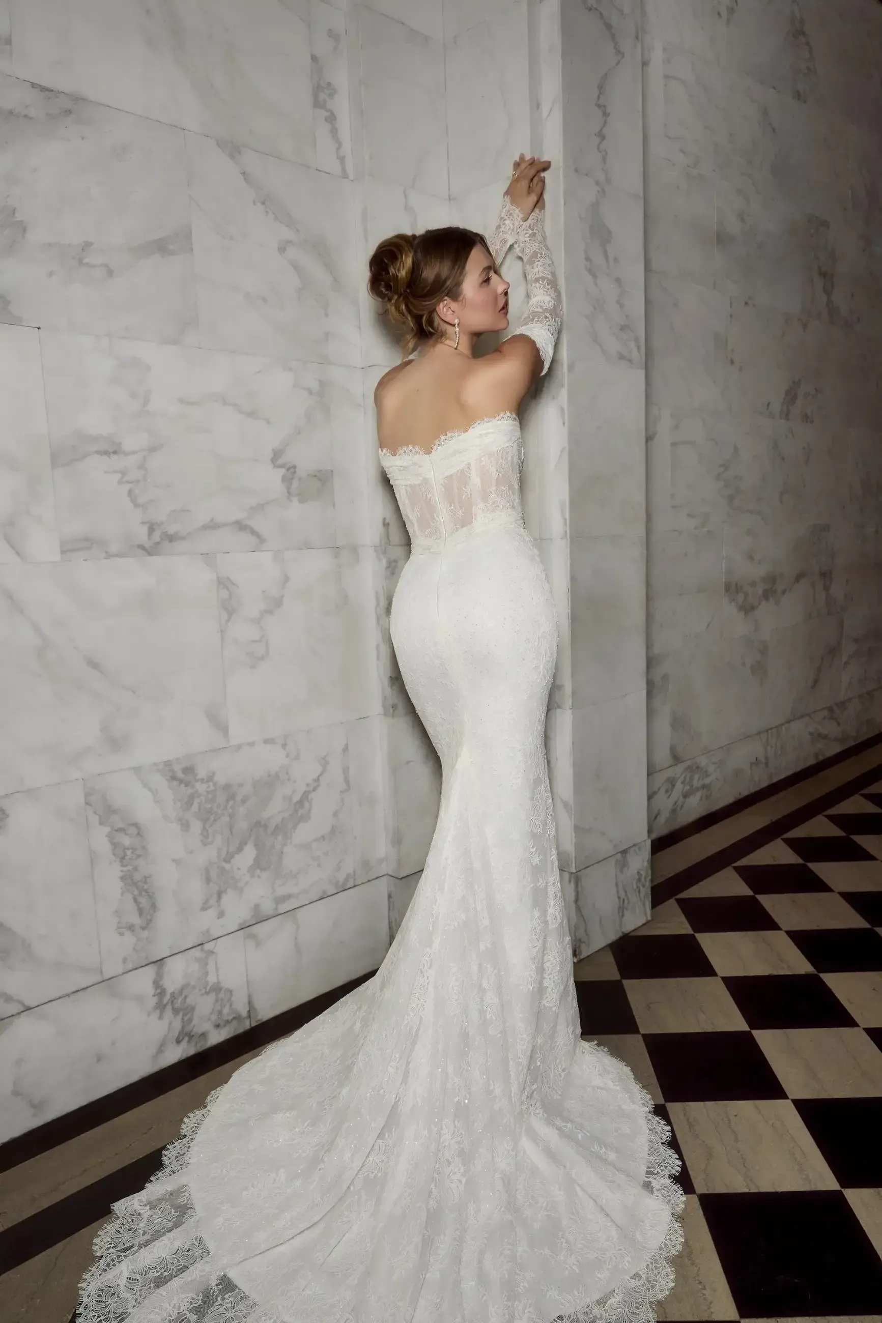 Woman in a white off-the-shoulder wedding gown poses against a marble wall; long train and sleeves.