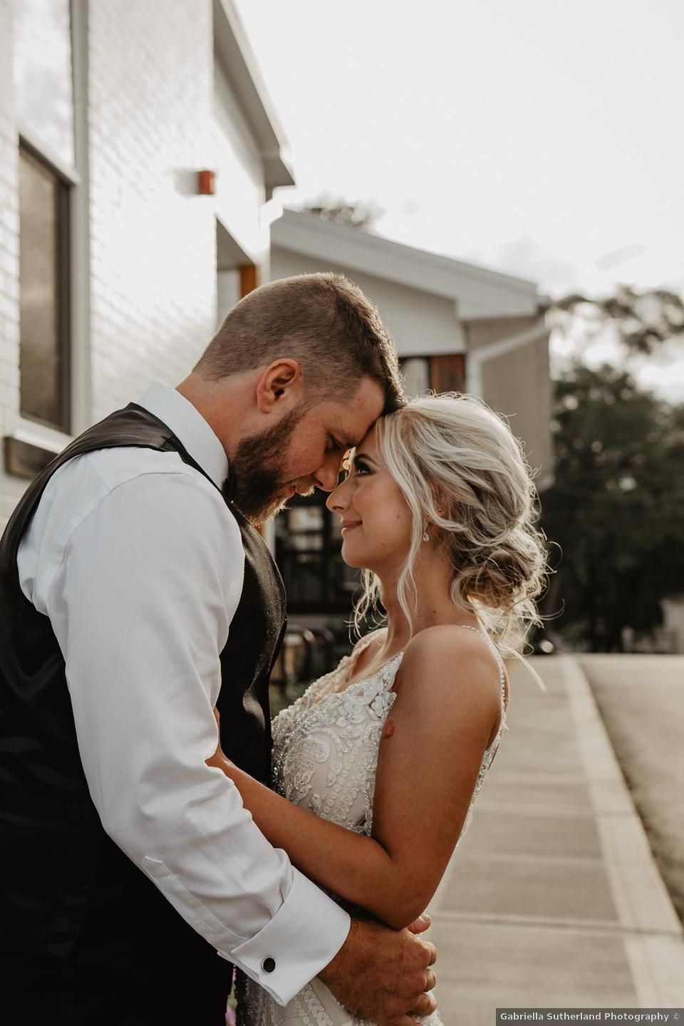 Wedding couple embraces, foreheads touching, outside near a building.