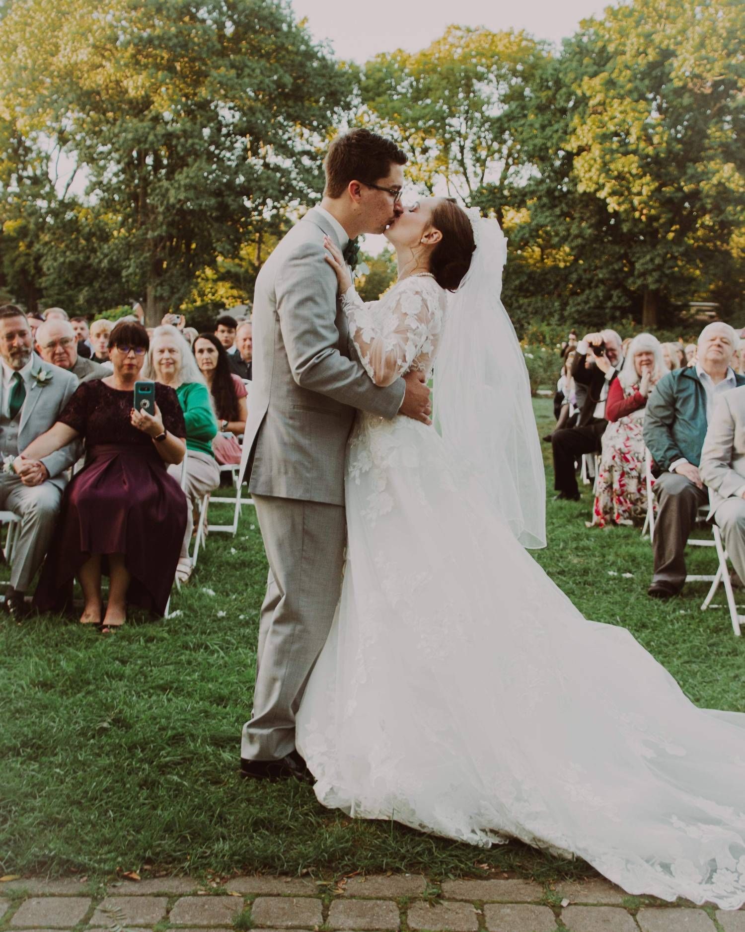 Couple kissing at outdoor wedding ceremony. Bride in white dress, groom in gray suit. Guests seated on lawn.