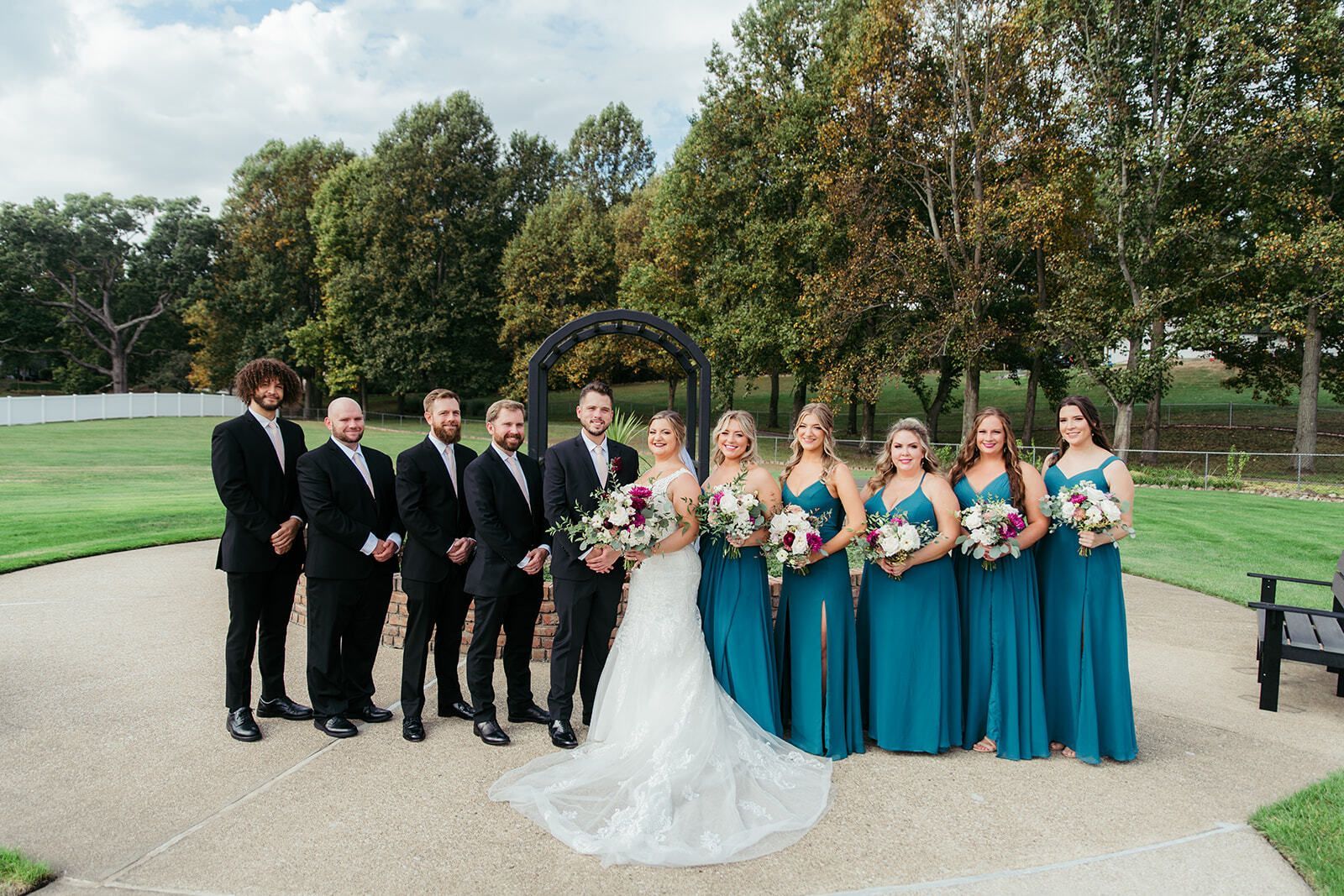 Wedding party poses outdoors; bride in white gown, bridesmaids in teal dresses, groomsmen in black suits.