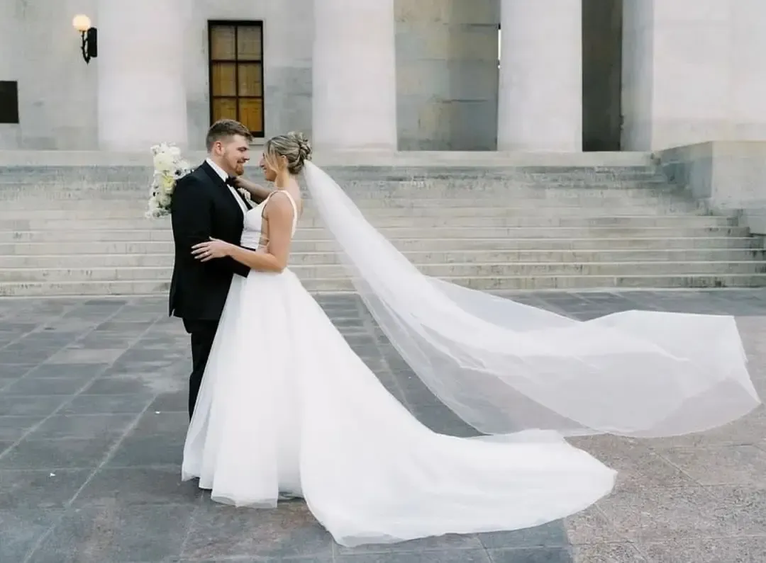 Bride and groom embrace in front of a building with steps. The bride wears a white dress with a long veil. The groom wears a black suit.