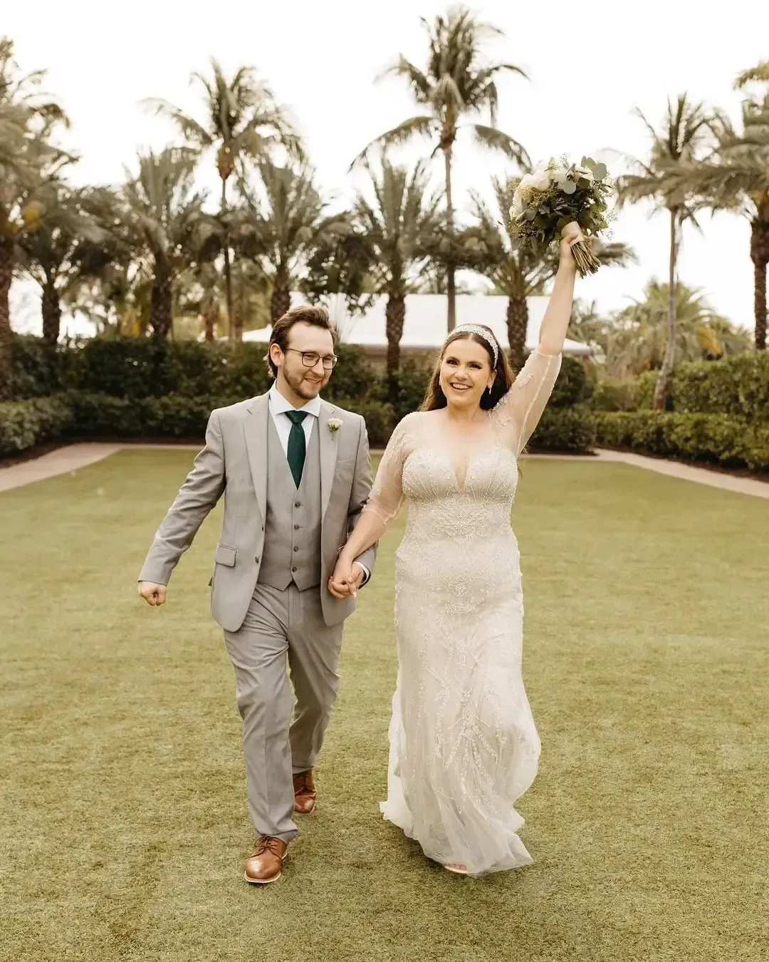 Newlyweds holding hands, woman raising bouquet, walking on grass. Palm trees in background.