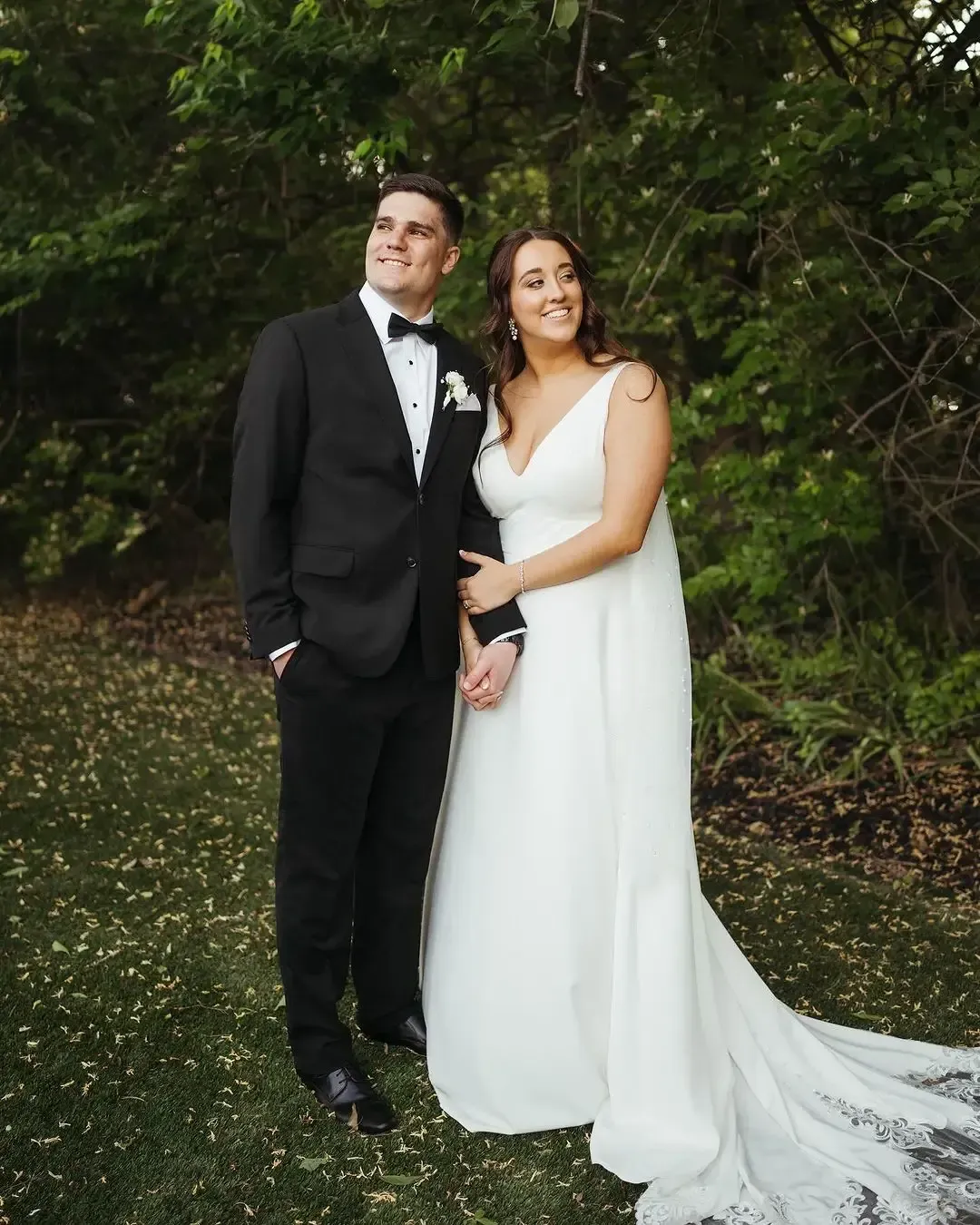 Couple in wedding attire, holding hands outdoors; man in tuxedo, woman in white gown.