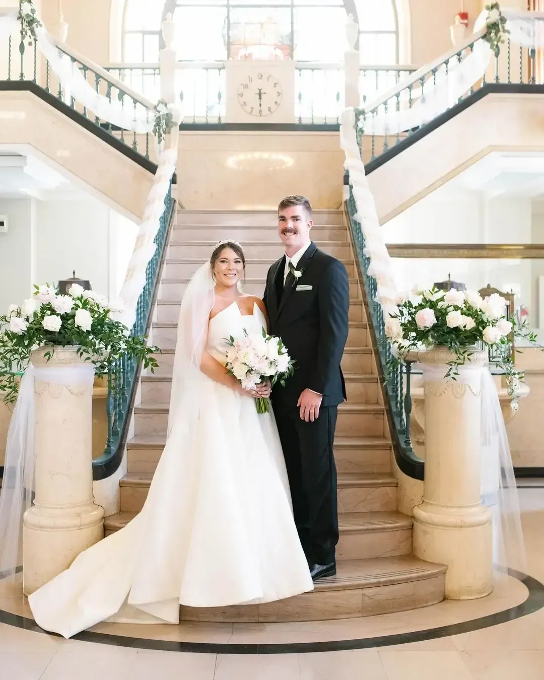 Bride and groom on a grand staircase, smiling. Bride holds bouquet, wearing veil and strapless gown. Groom in a suit.