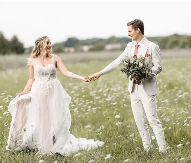 Bride and groom holding hands in a field of flowers; the bride smiles at the groom.