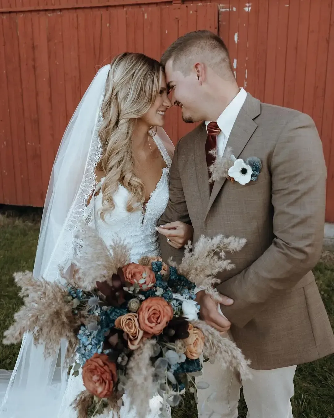 Wedding couple, bride in white dress and veil, groom in tan suit, looking at each other. Rustic setting, holding bouquet.