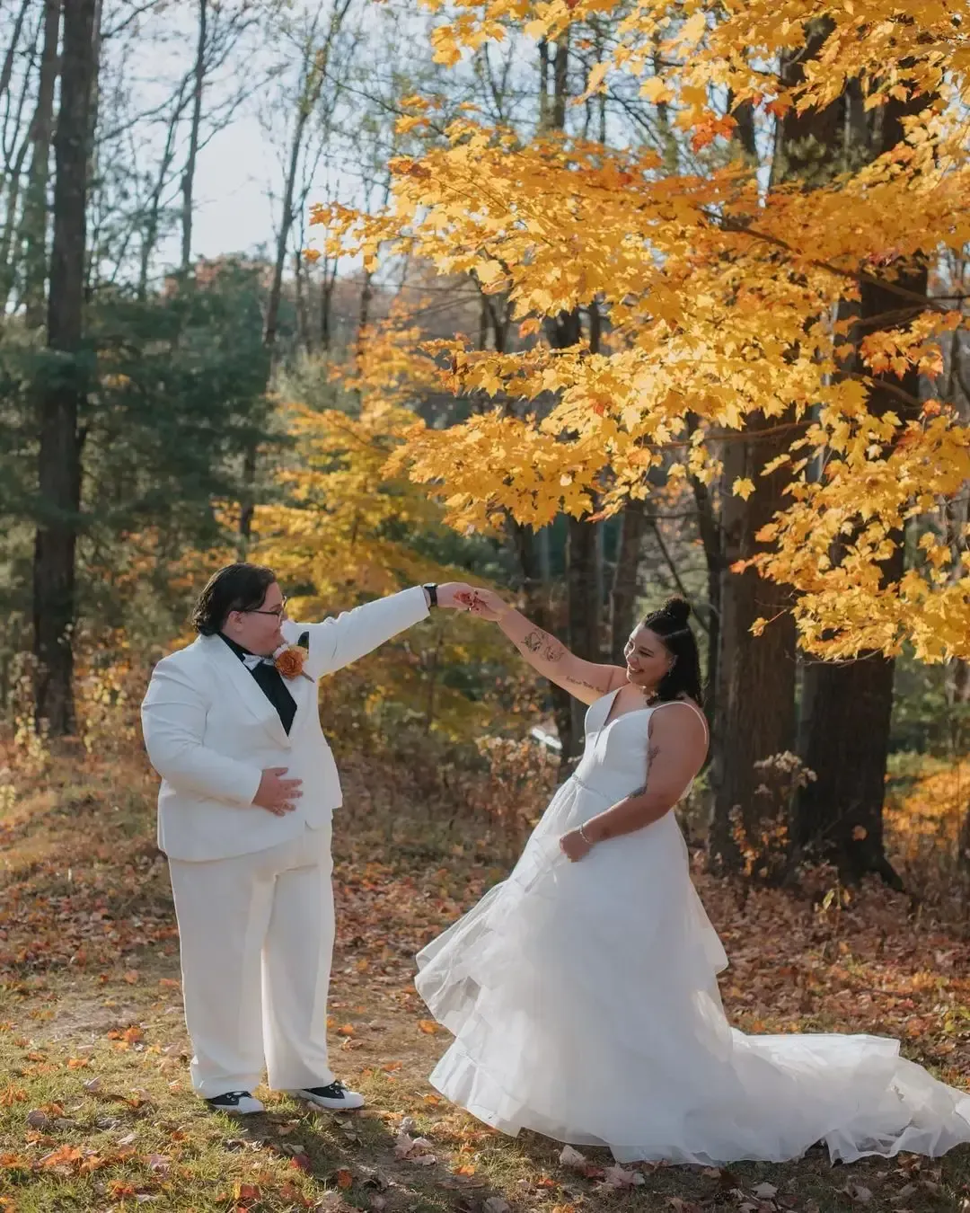 Two people in wedding attire dance beneath fall foliage.