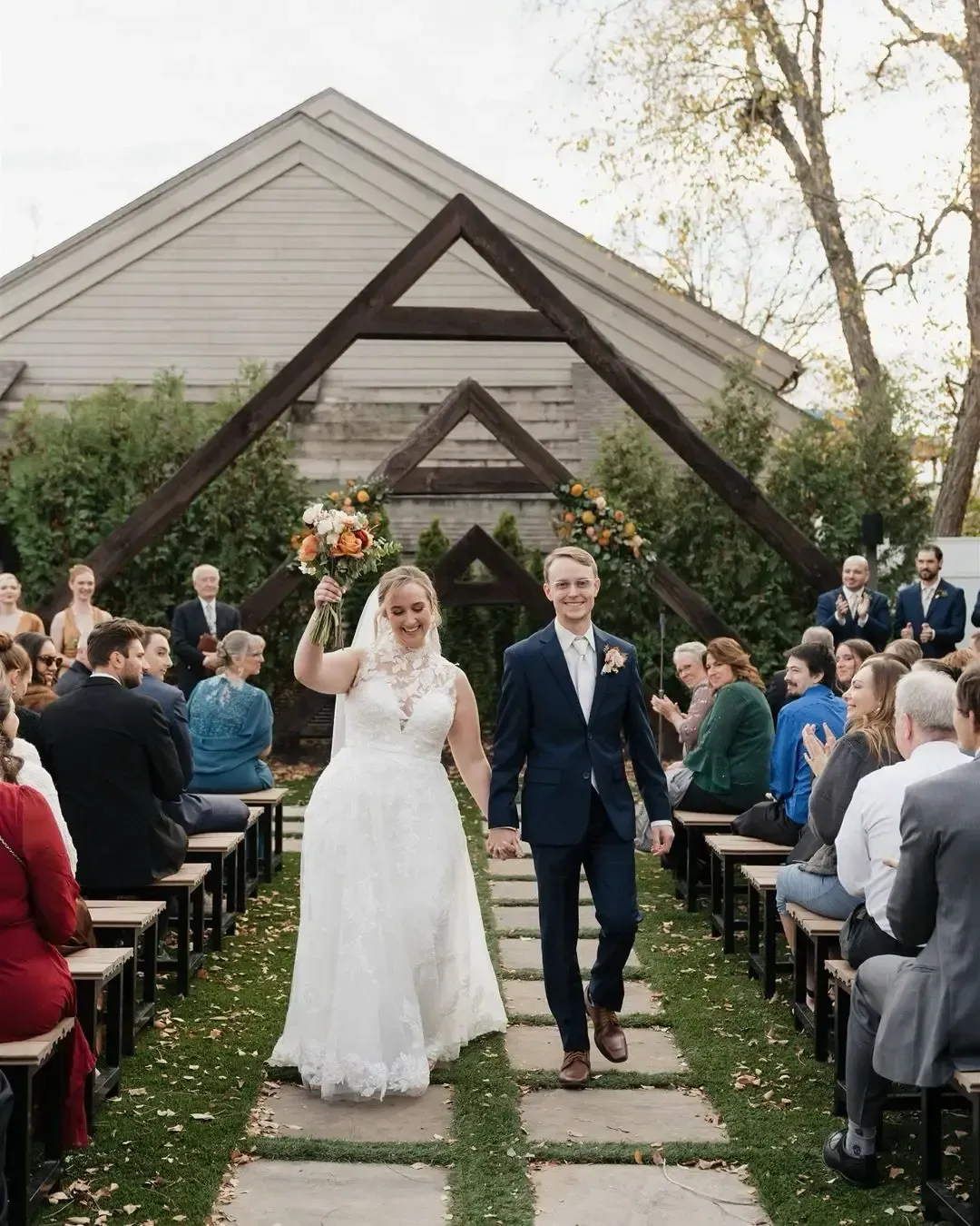 Newly married couple walking down aisle, holding hands. Bride raises hand, smiling, groom smiles. Outdoor wedding.