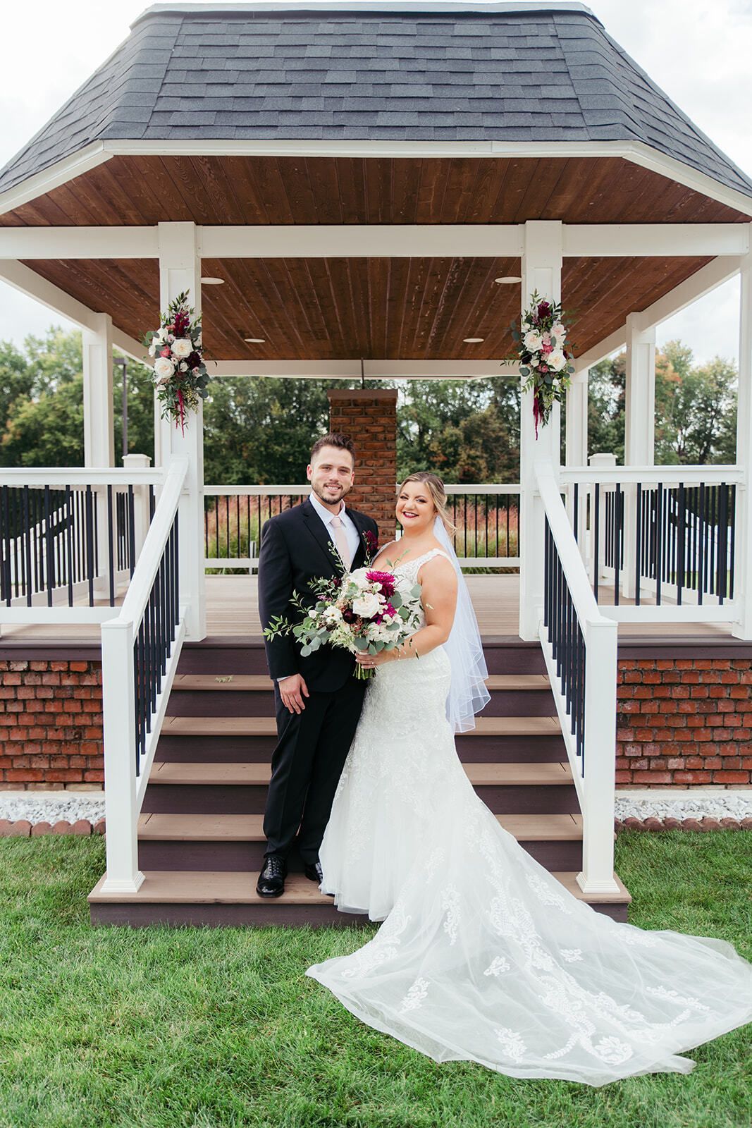 Couple in wedding attire posing on steps of gazebo, holding bouquet.