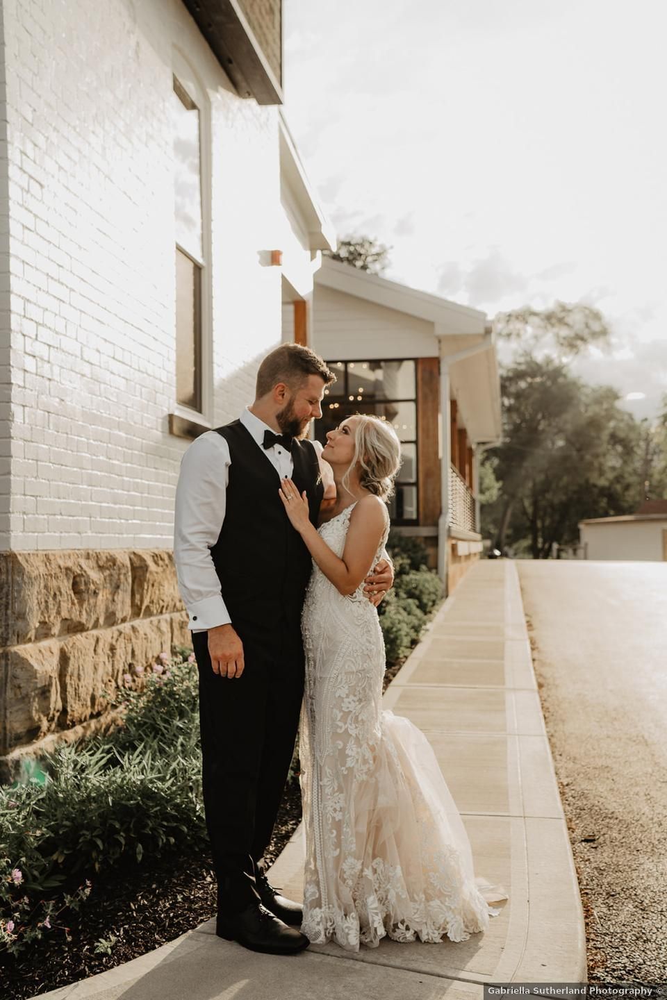 Bride and groom embrace outside building. Bride wears white lace dress, groom a black vest.