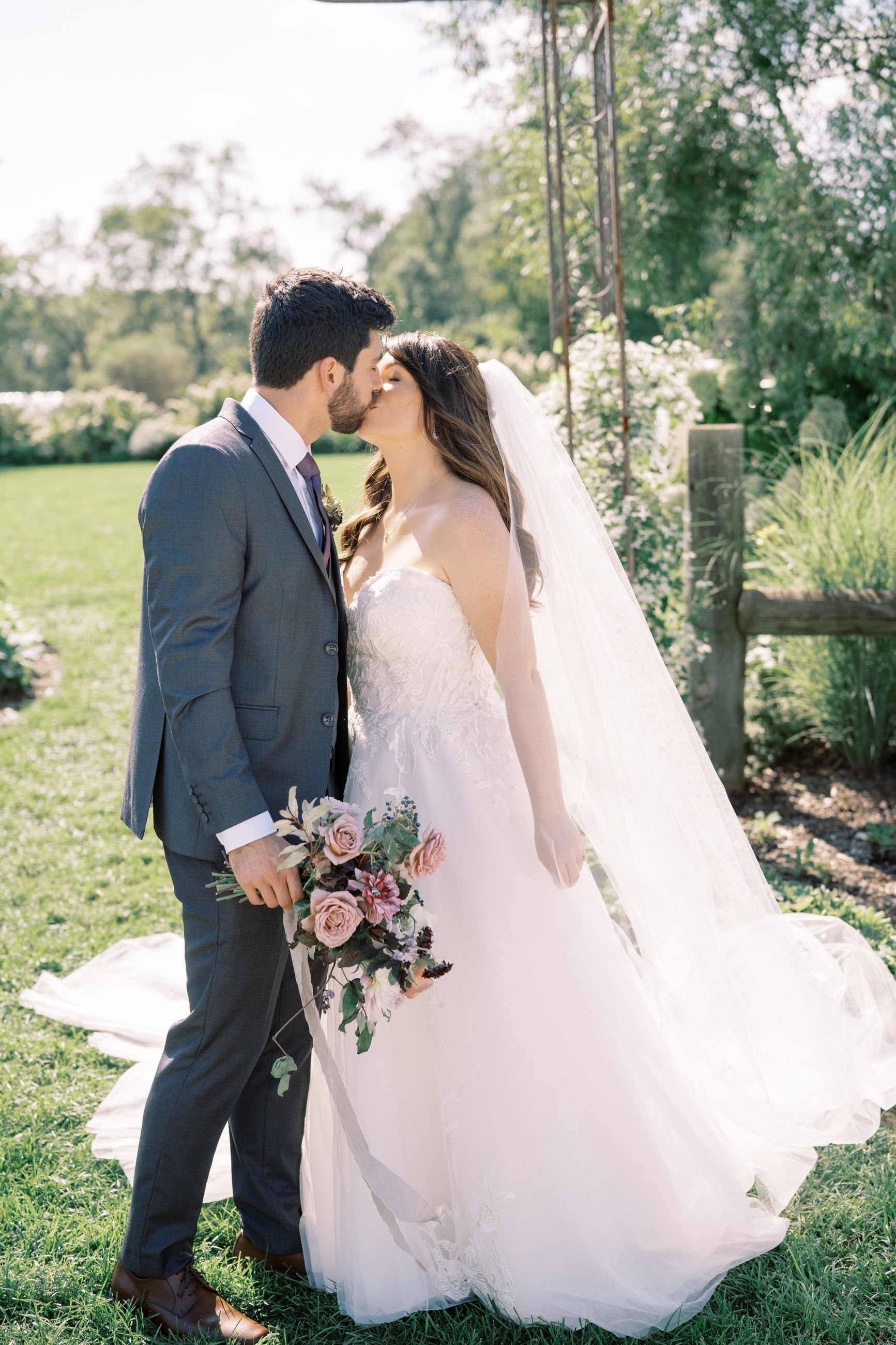 Couple kissing, bride holding bouquet, groom in suit; sunny outdoor wedding.